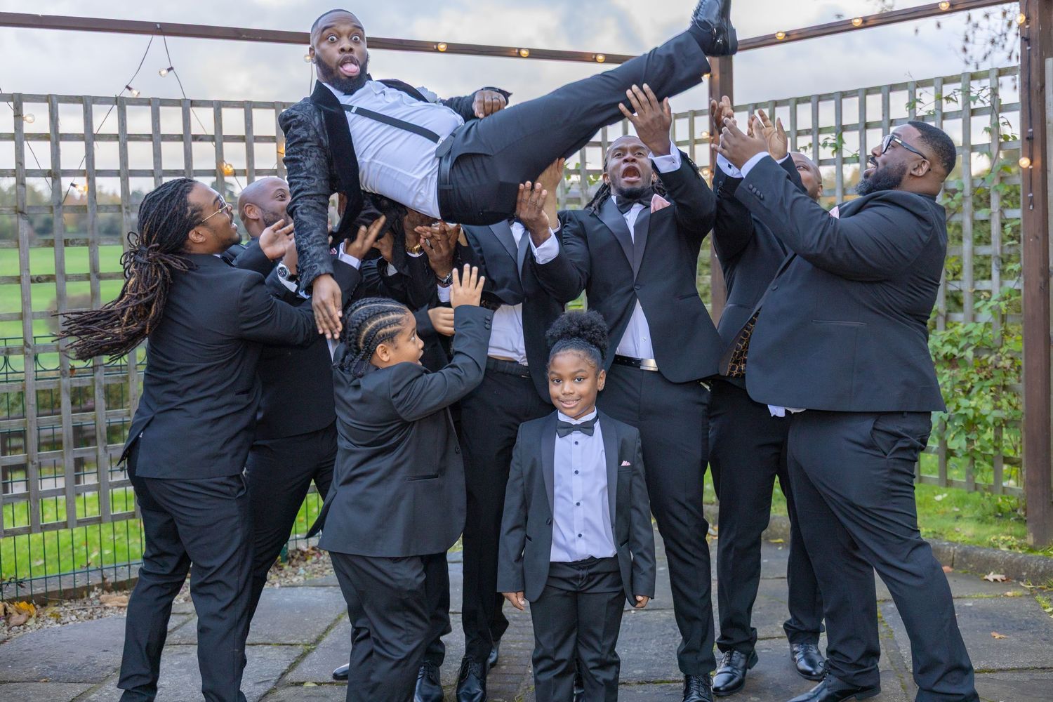 Groomsmen in black suits lift the groom high while two young boys pose in front at an outdoor wedding.