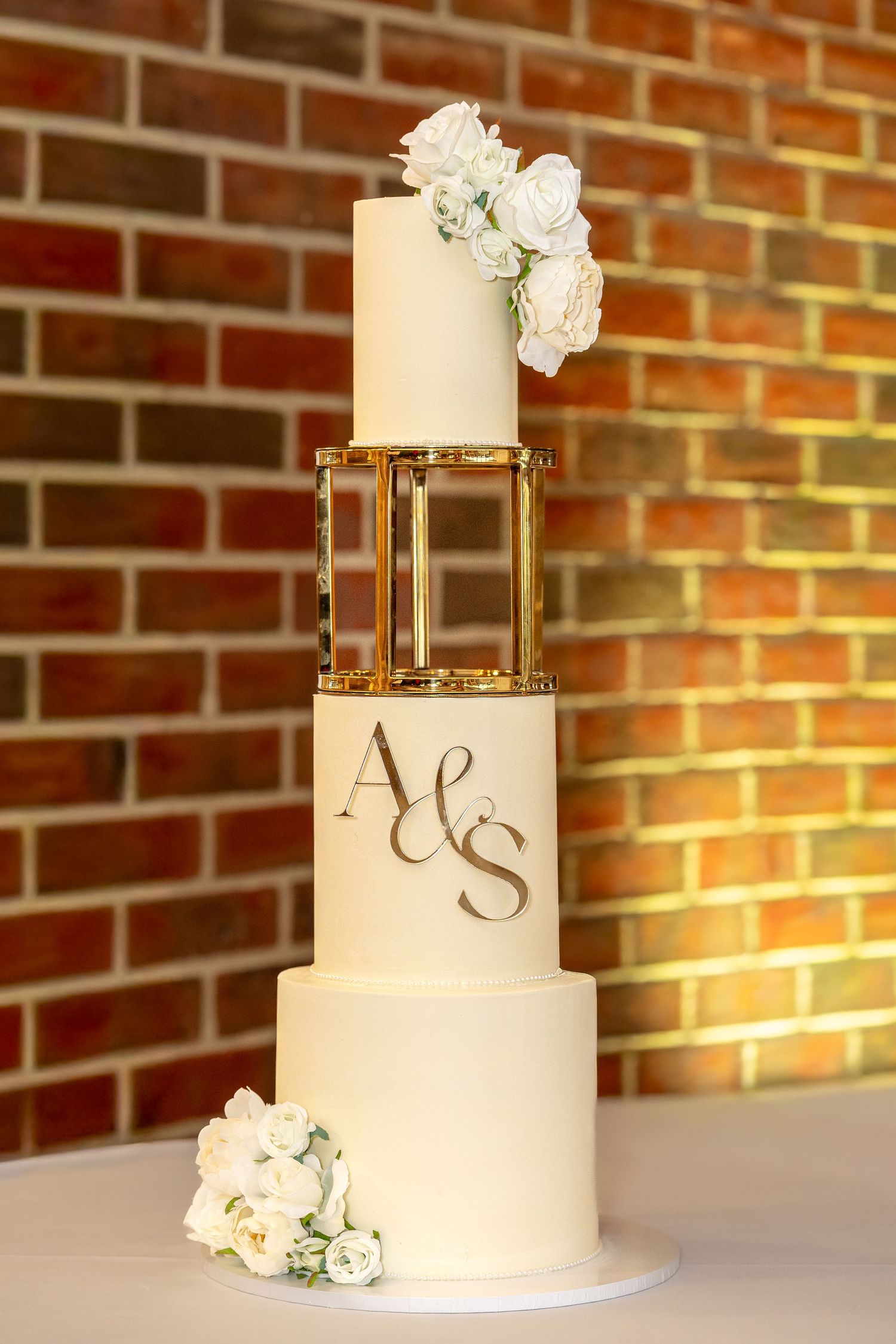 Elegant three-tier white wedding cake with gold monogram A&S and white roses on brick background.