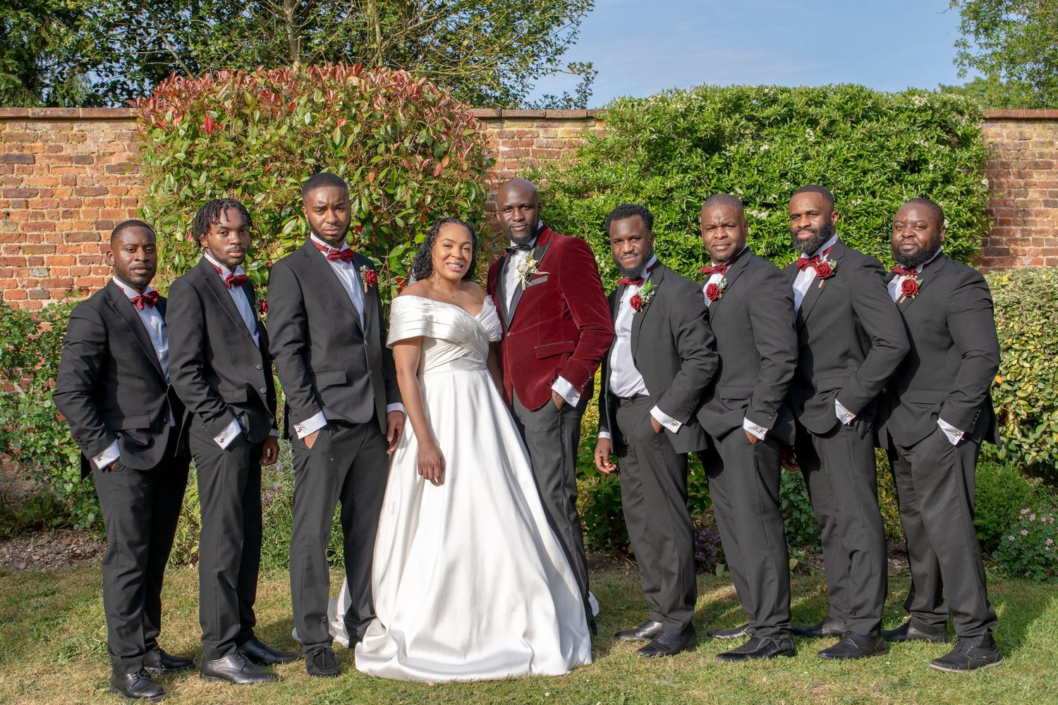 Bride in white gown and groom in red velvet suit pose with groomsmen in black tuxedos outdoors.