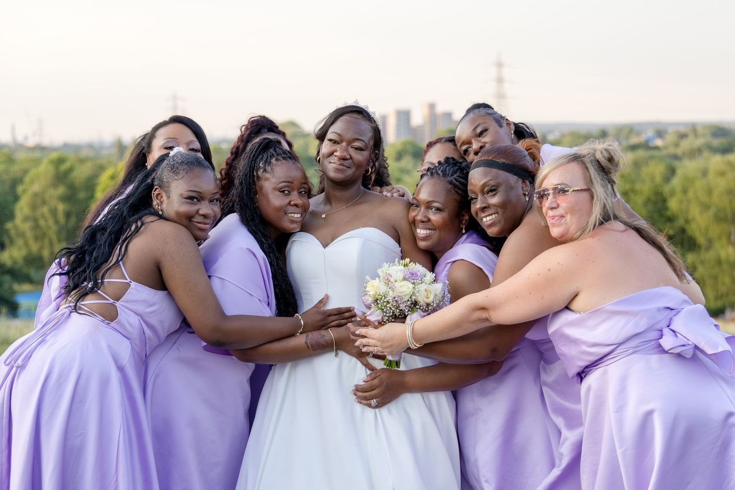 Bride holding bouquet surrounded by smiling bridesmaids in lavender dresses in a warm group hug.