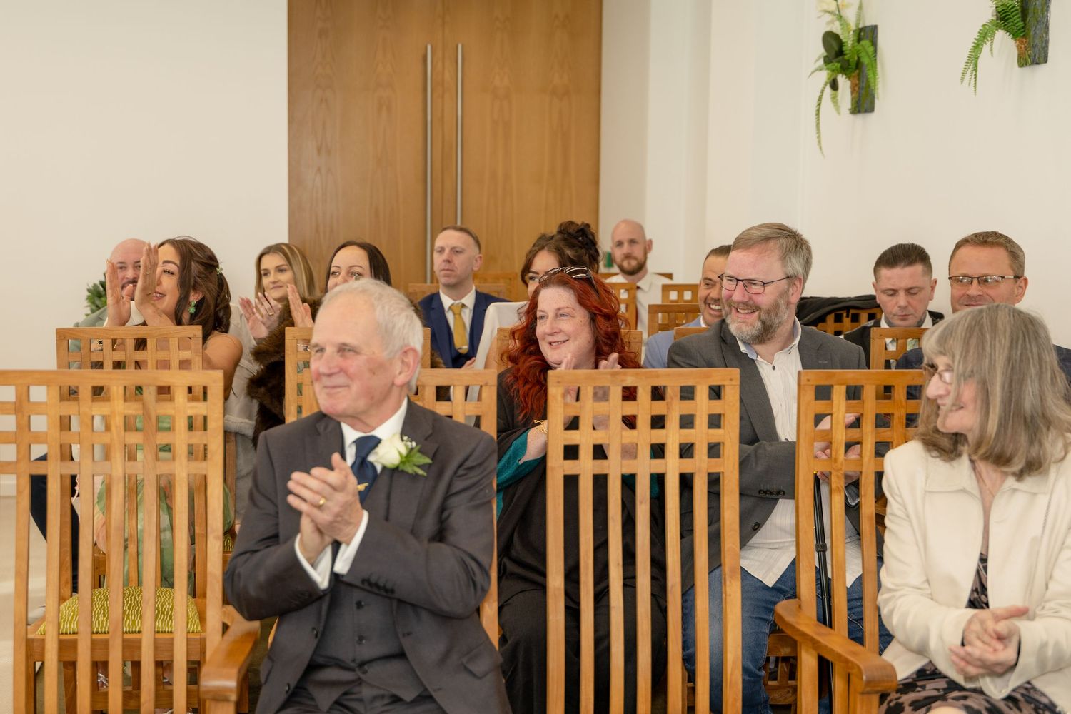Wedding guests applaud and smile while seated in wooden pews during ceremony.