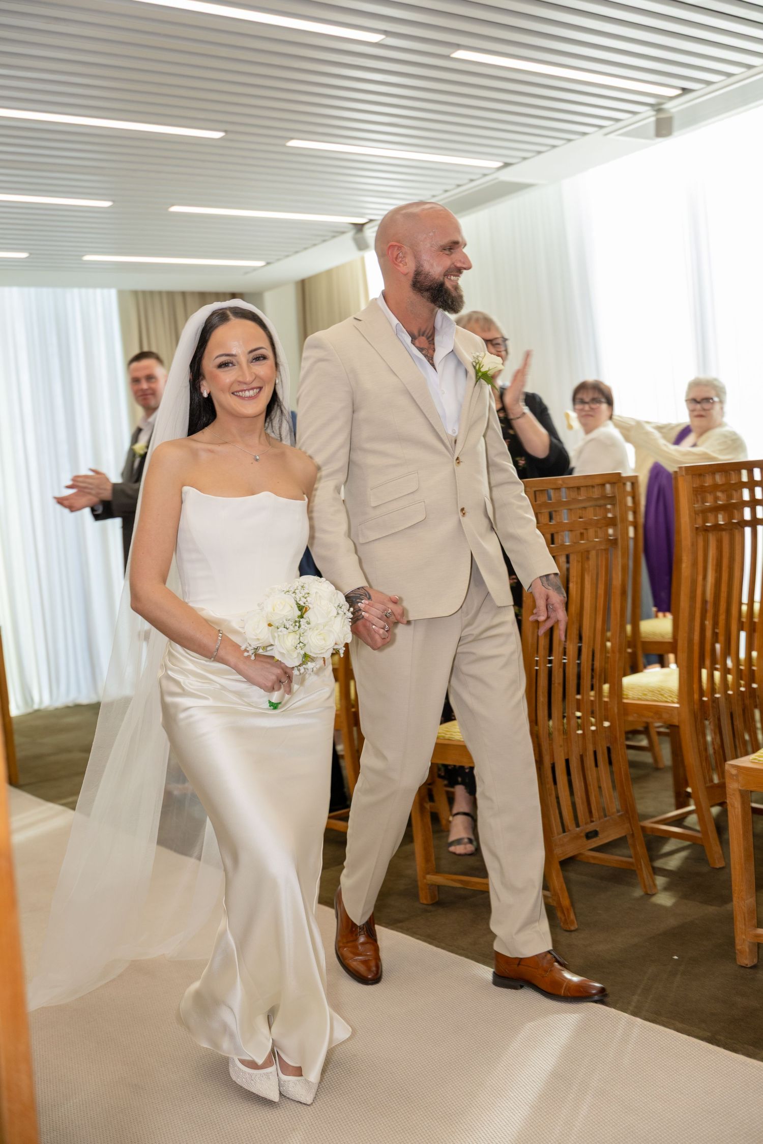 Smiling bride and groom walk hand in hand down the aisle past seated wedding guests.