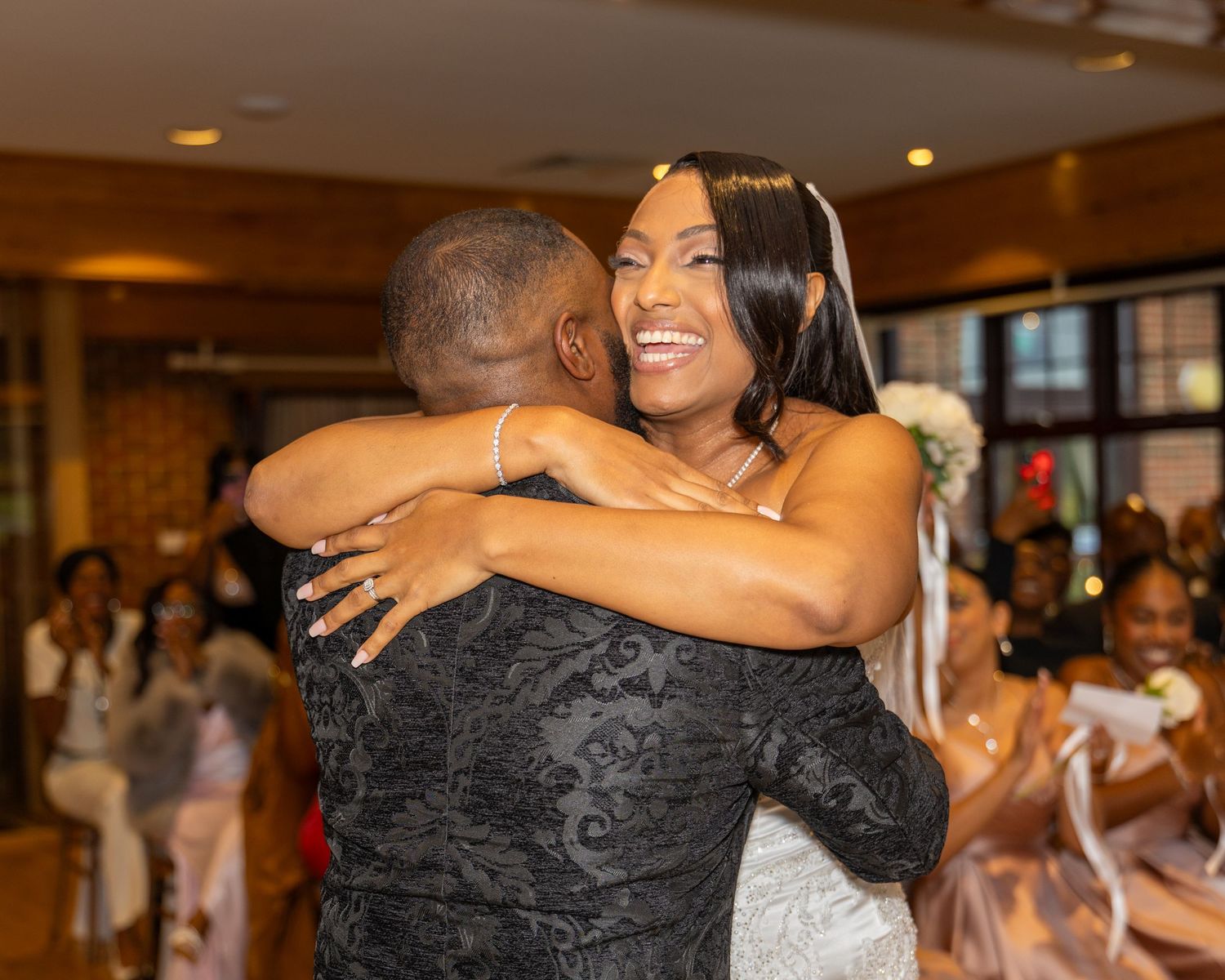 A joyful bride in a white strapless gown and veil shares a warm embrace with a guest at her wedding reception.