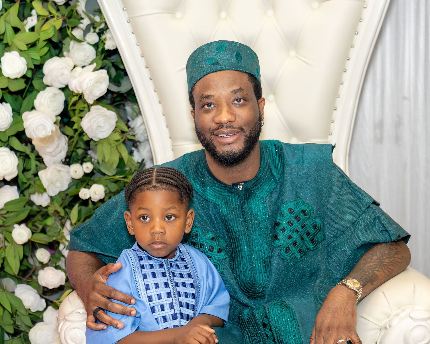 Man and young boy in matching green traditional African attire pose together on a white throne chair.