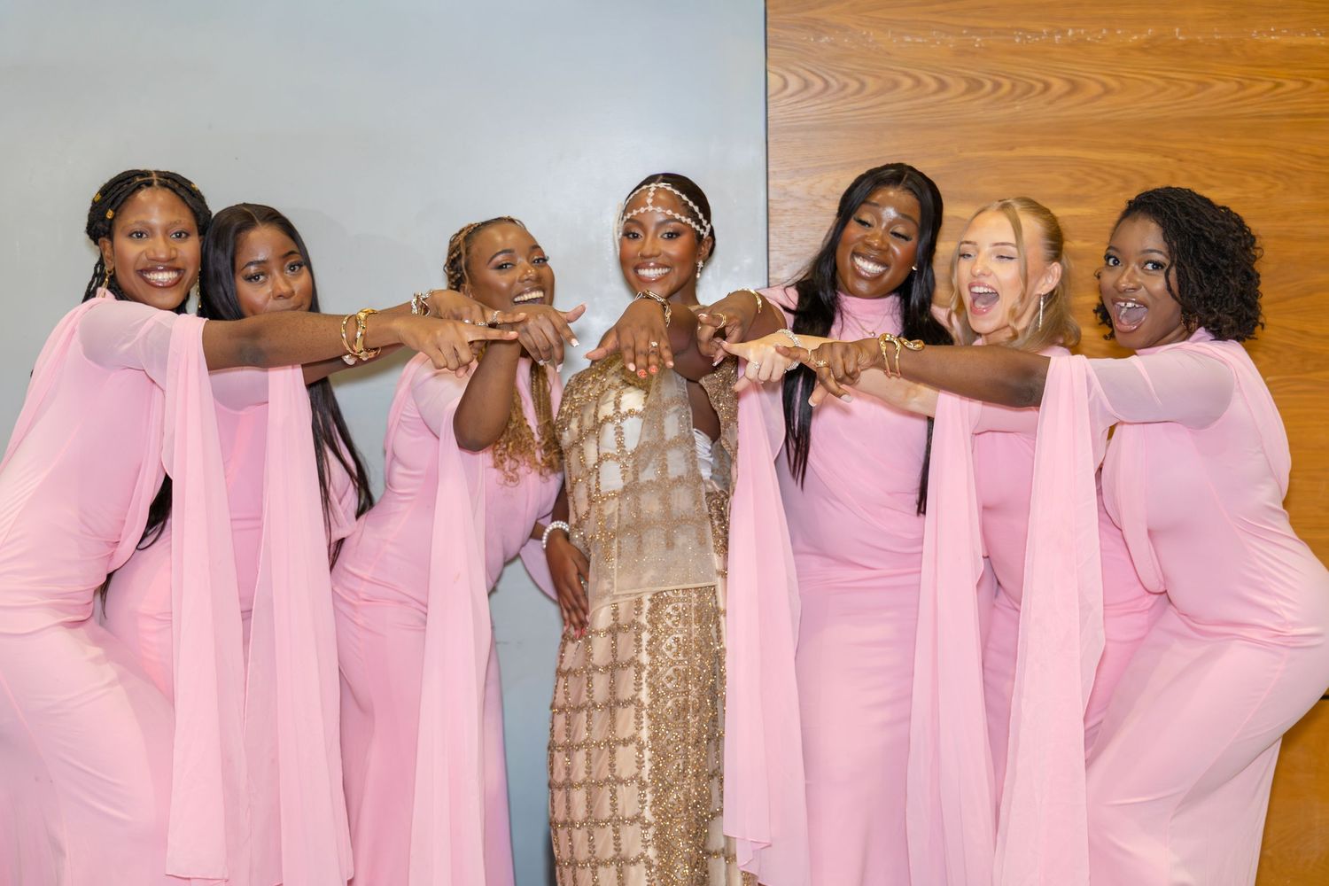 Bride in gold beaded dress celebrates with six bridesmaids in pink dresses pointing at her ring.
