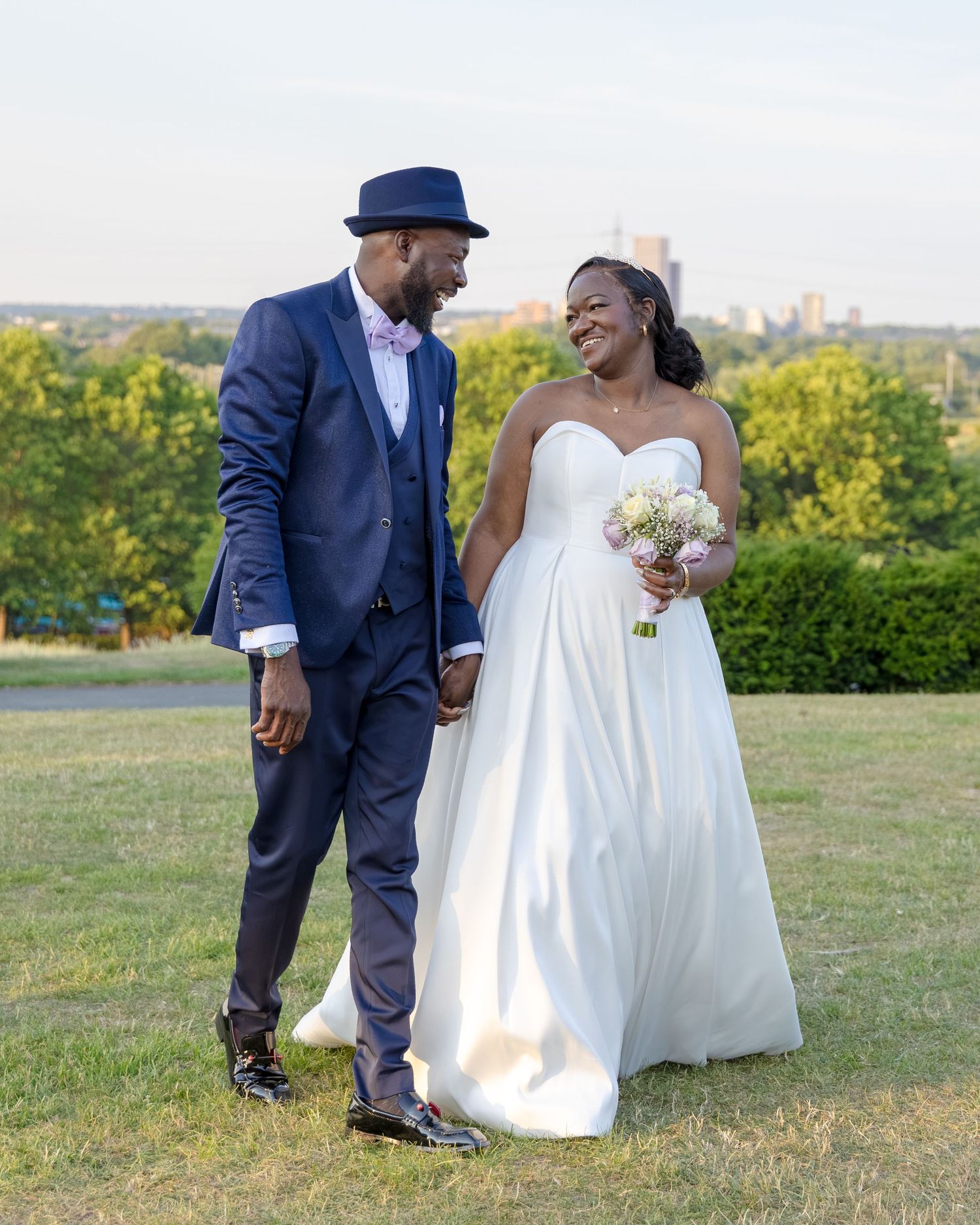 A joyful newlywed couple in wedding attire hold hands on a grassy hill with a city skyline backdrop.