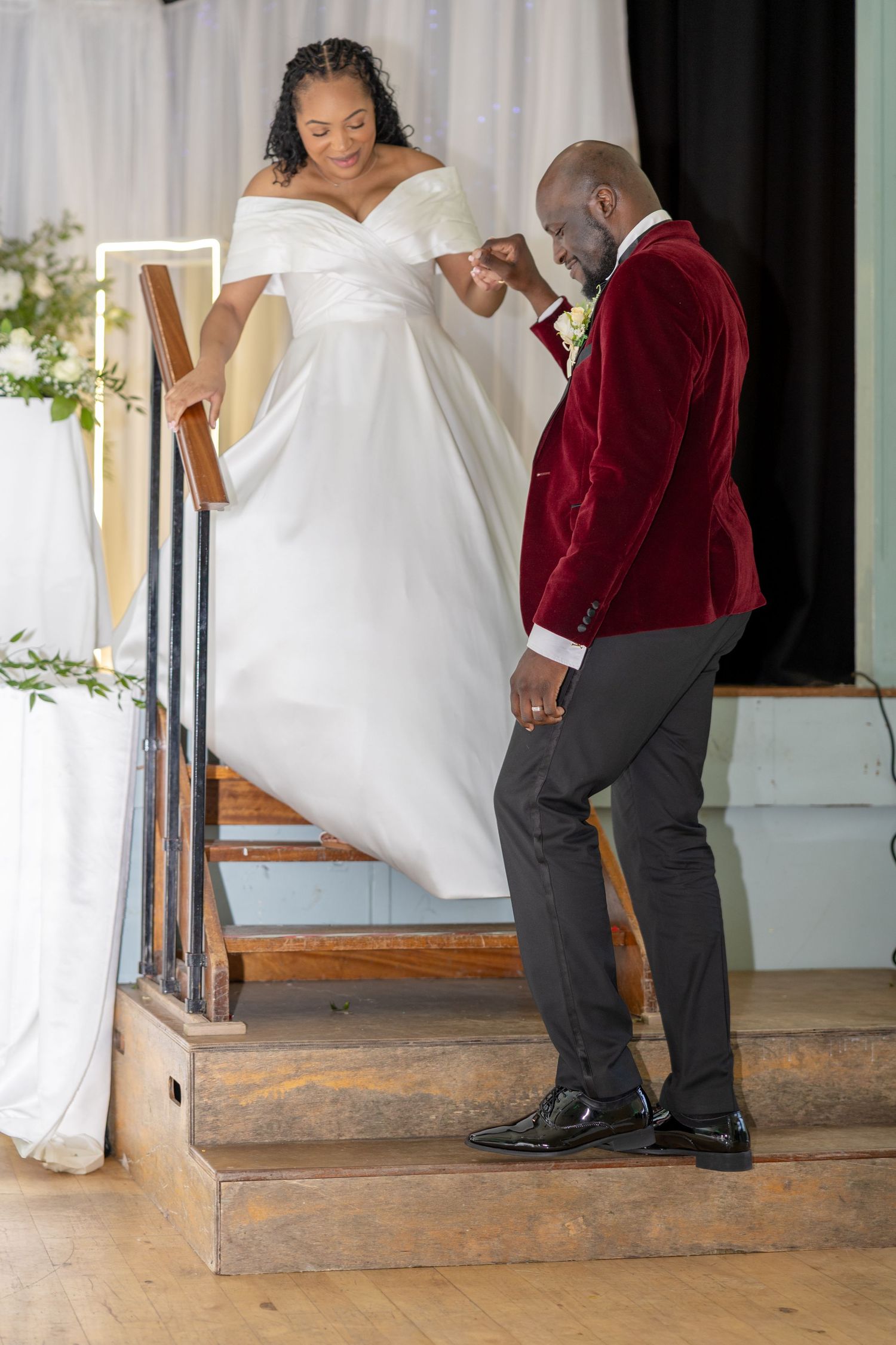 Groom in red velvet blazer kisses bride's hand as she descends wooden stairs in elegant white wedding gown.