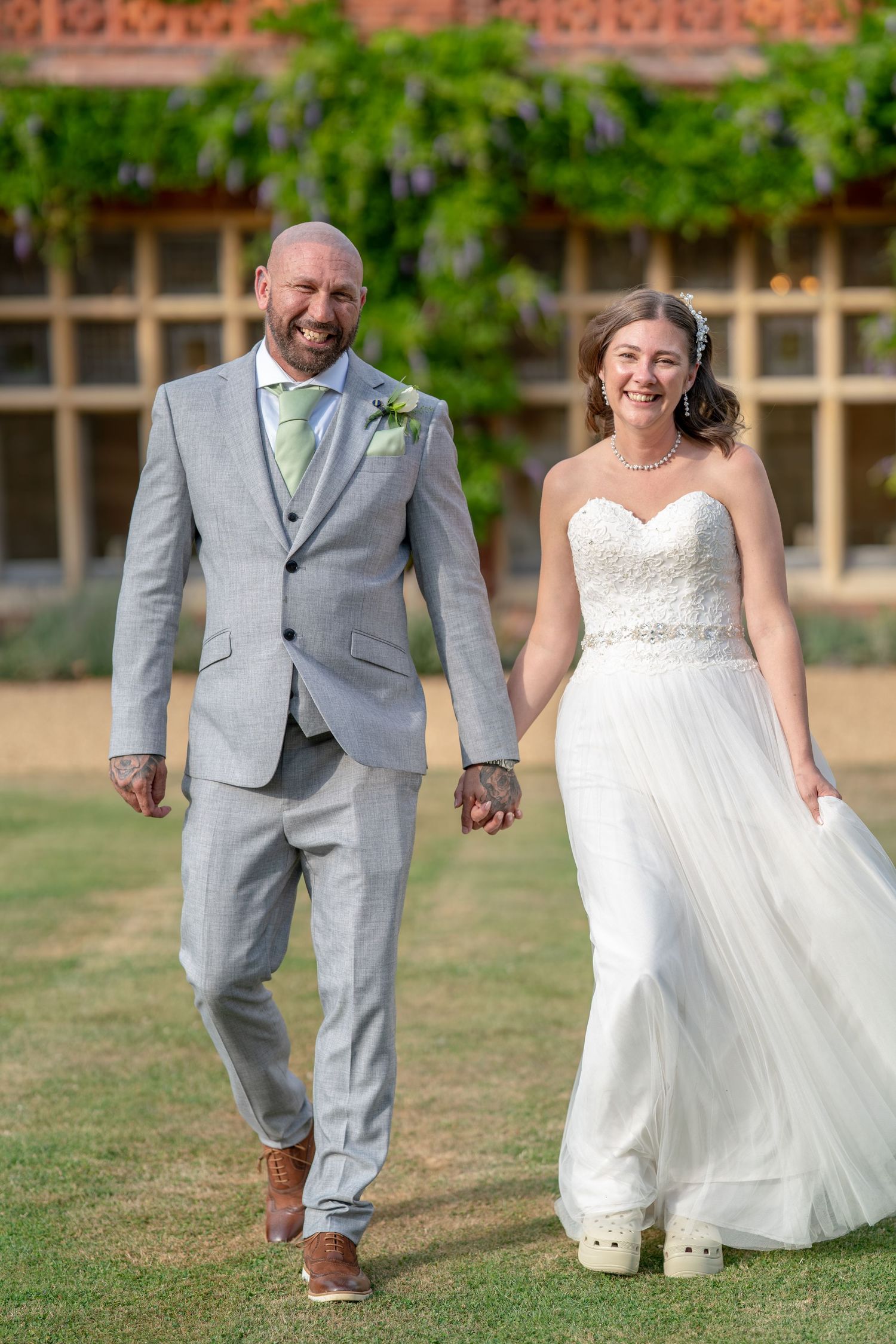 A smiling bride in a white strapless gown and groom in a gray suit hold hands while walking on grass at an outdoor wedding.