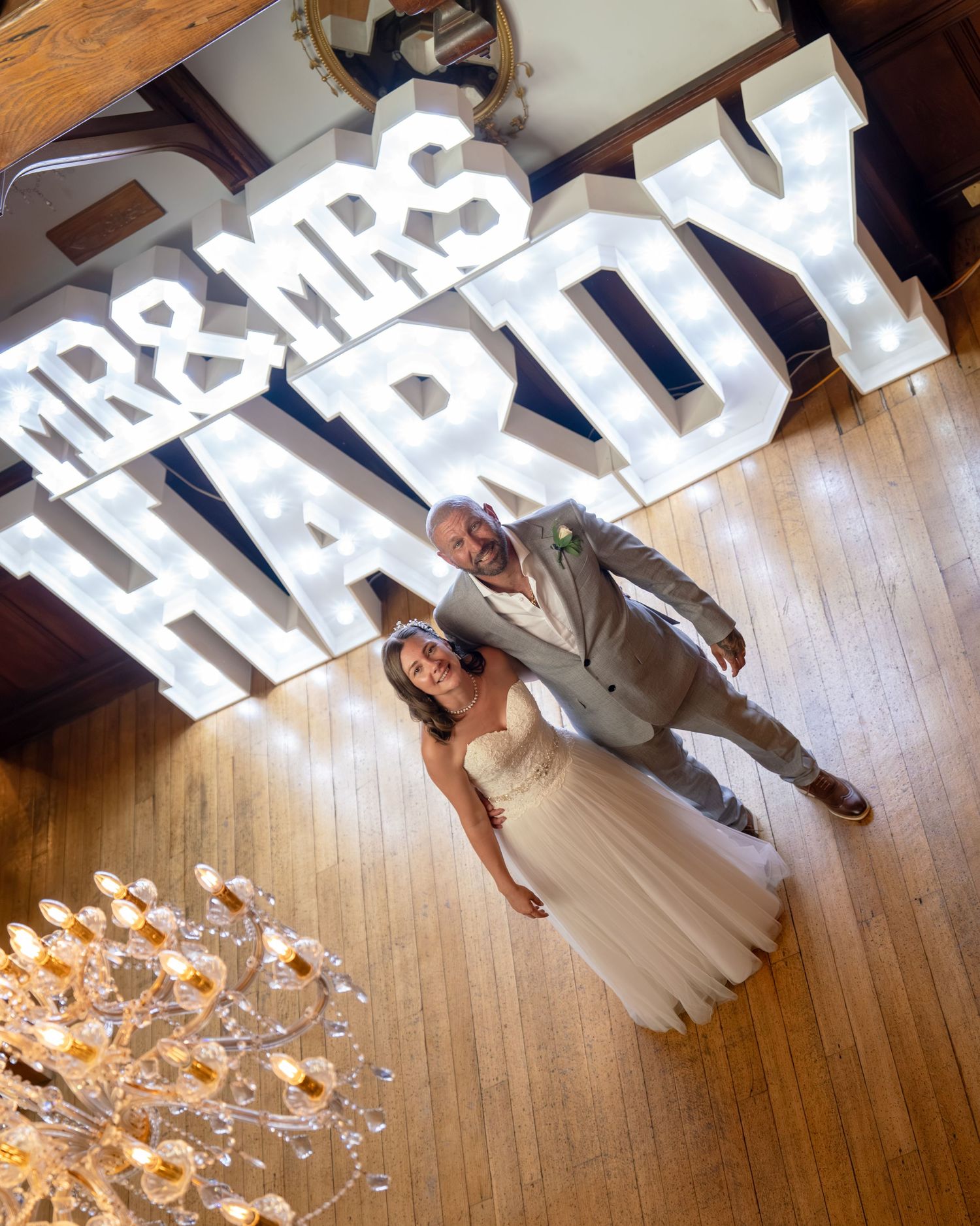 Bride and groom posing in front of illuminated Mr & Mrs Hardy marquee letters on a wooden dance floor.
