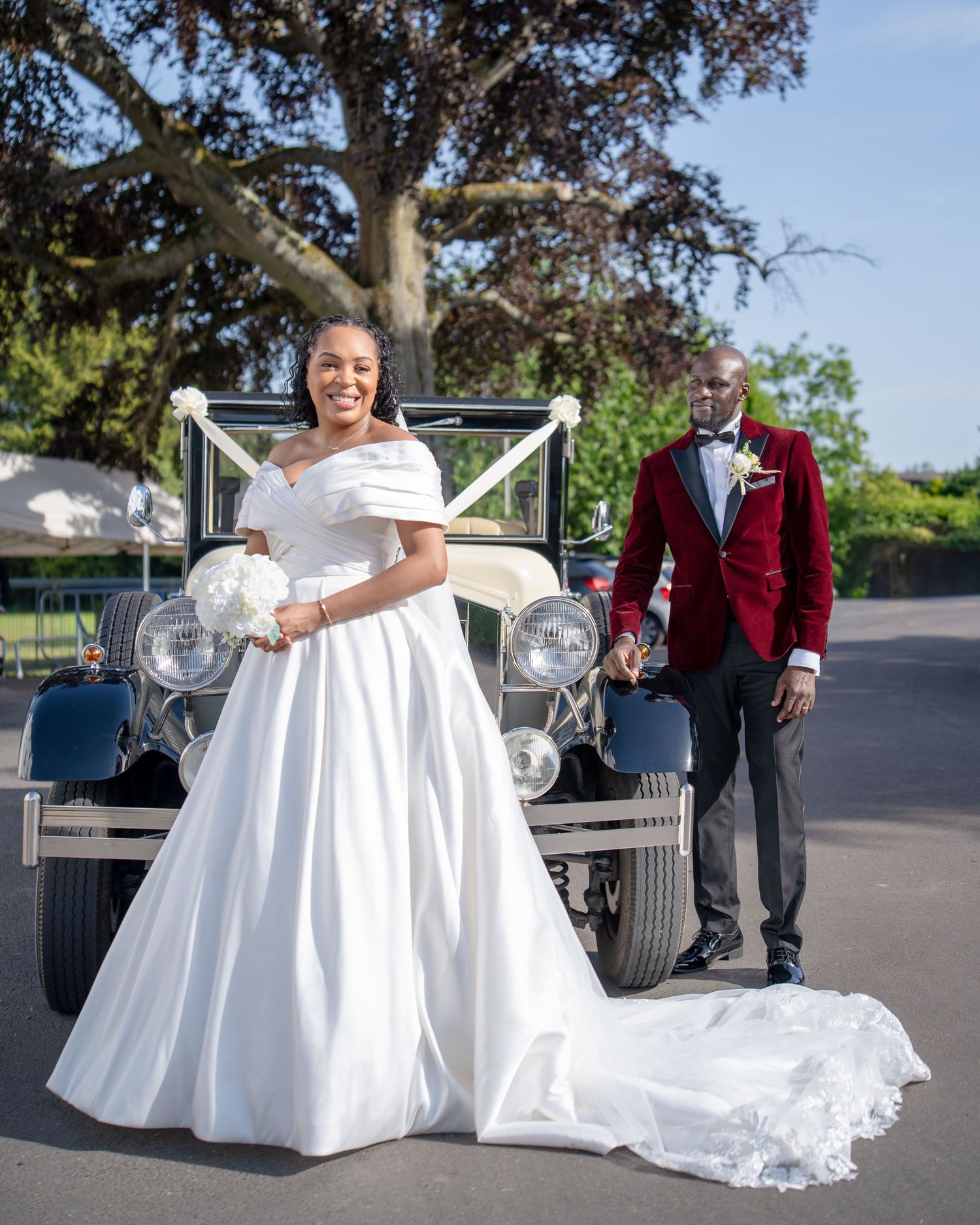 Bride in white off-shoulder gown and groom in red velvet tuxedo posing by a vintage classic car outdoors.