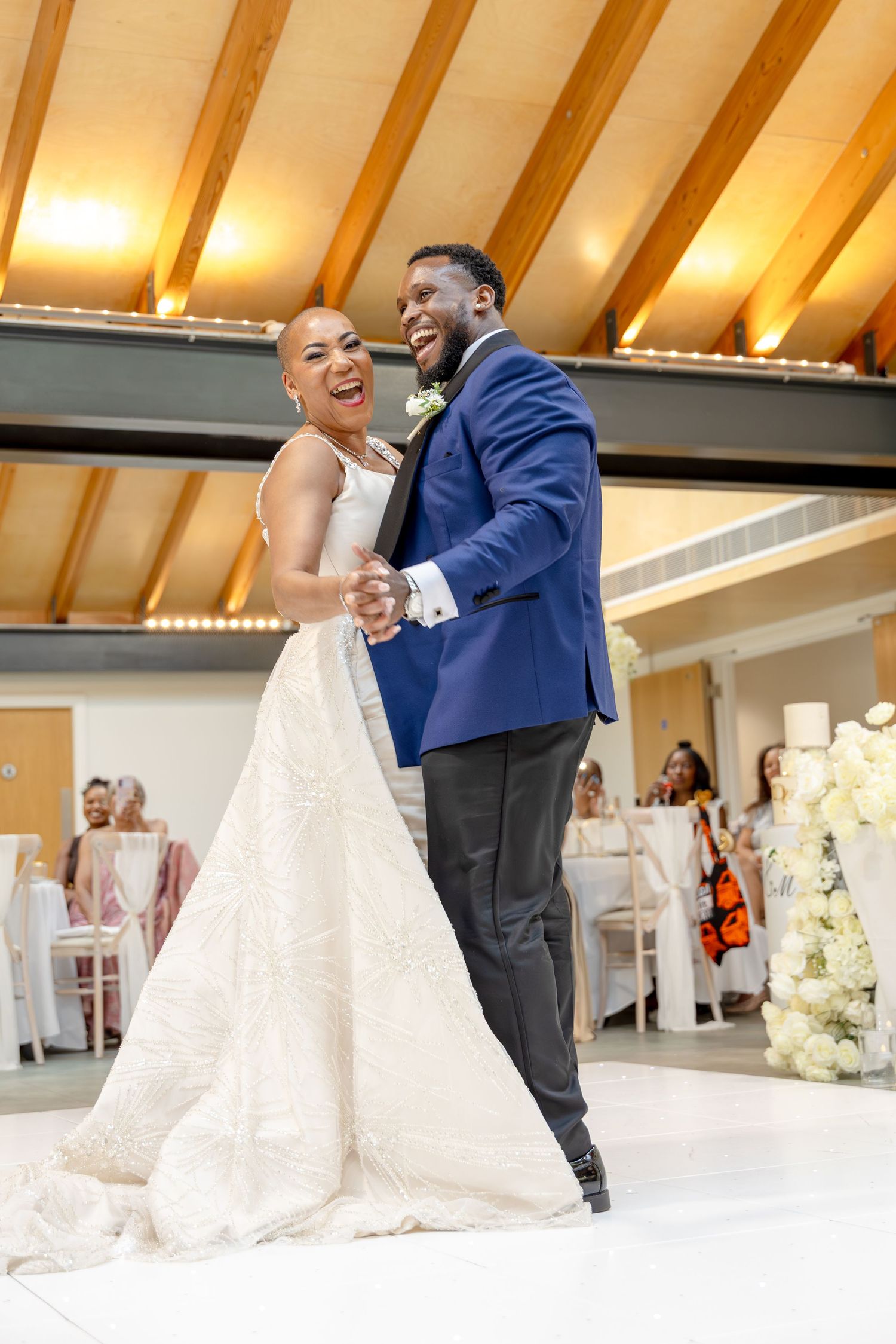 Joyful newlyweds share their first dance, bride in white gown and groom in blue suit, at elegant wedding reception.