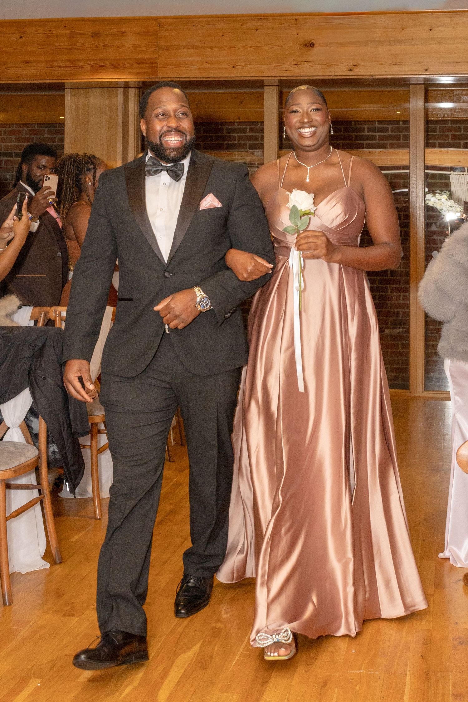 A groomsman in a black tuxedo escorts a bridesmaid in a rose gold satin gown holding a white rose at a wedding reception.