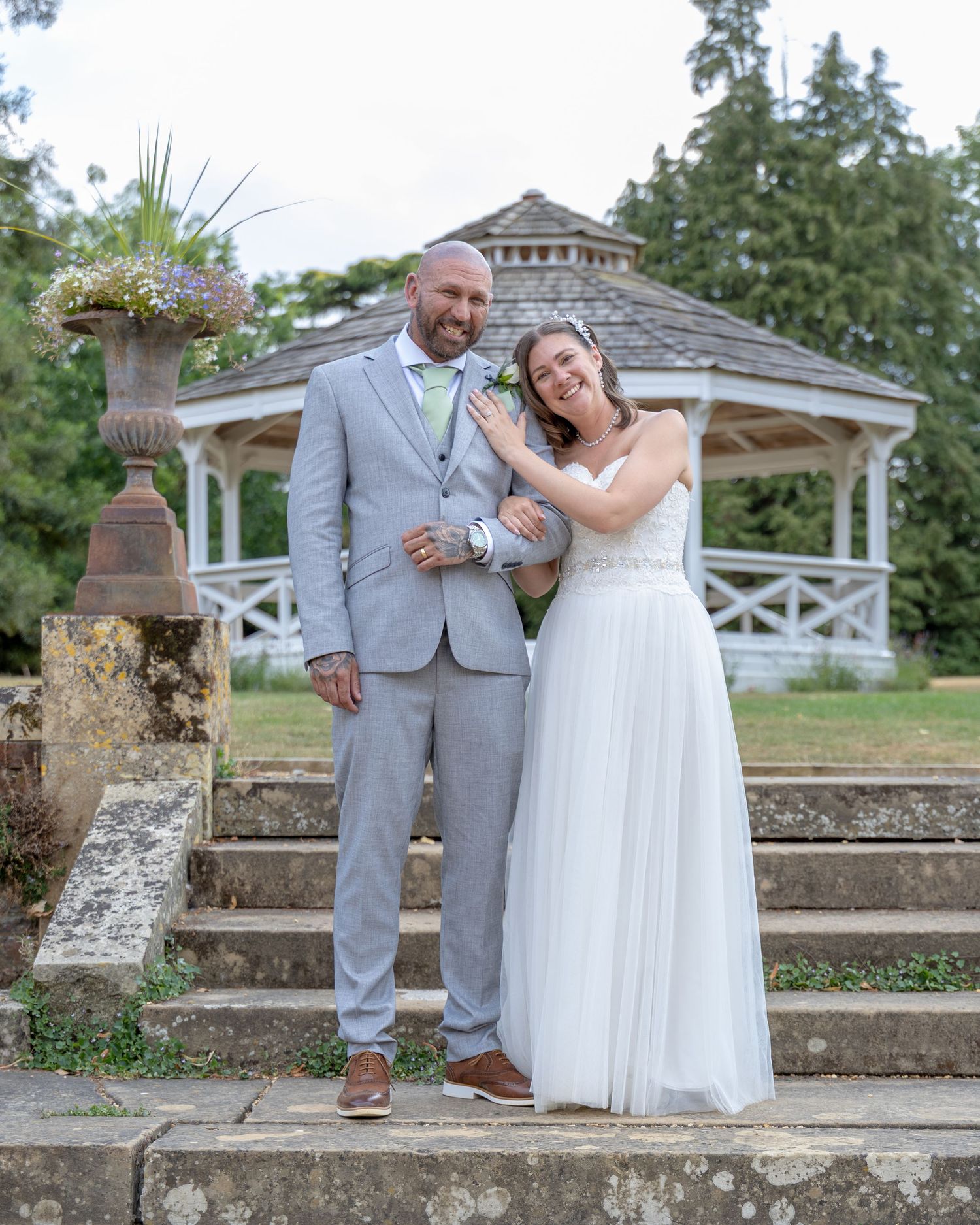 A couple poses on stone steps before a white gazebo, groom in gray suit, bride in white gown, at an outdoor wedding.