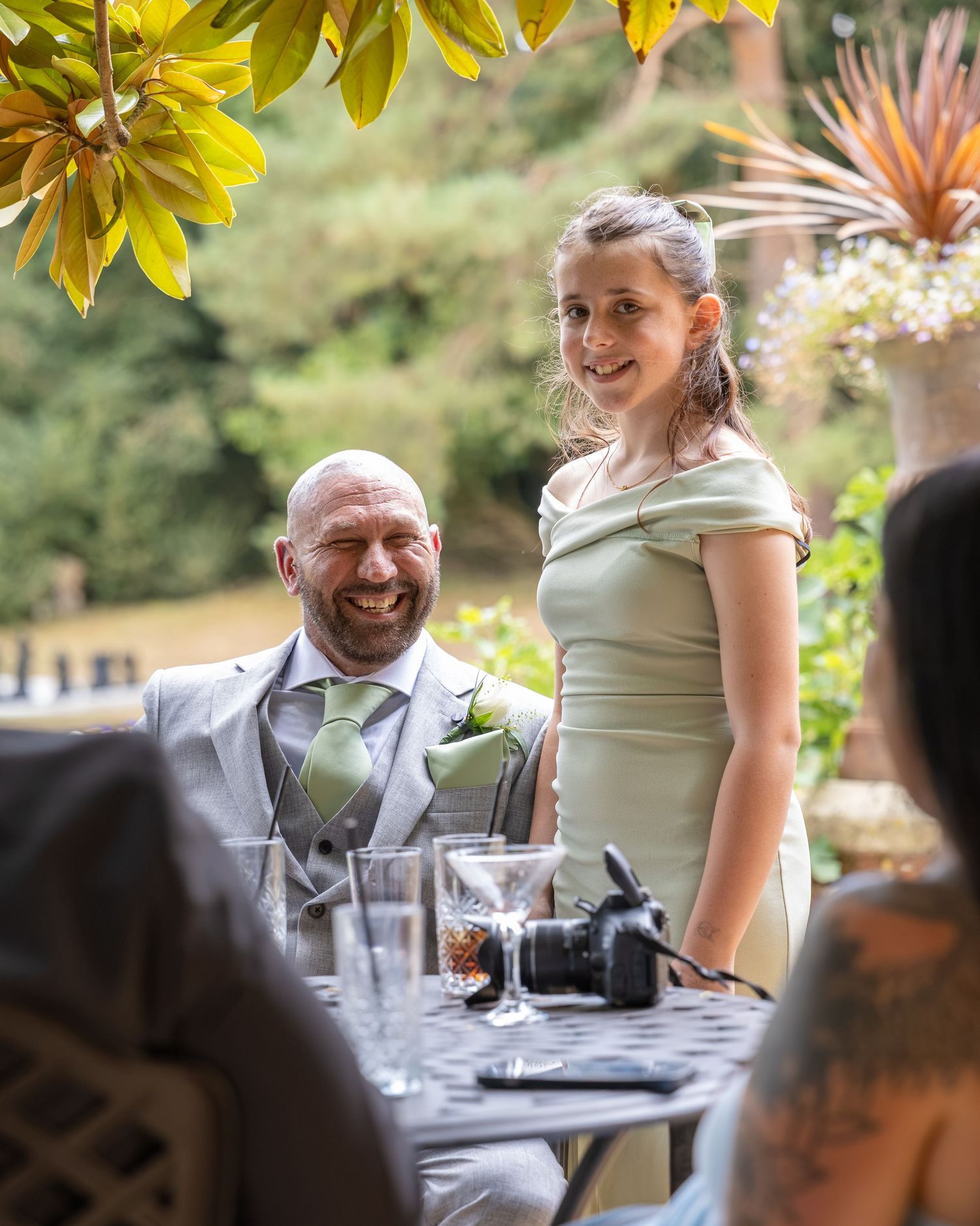 A smiling bald groom in a grey suit sits beside a woman in a sage green dress at an outdoor wedding reception.