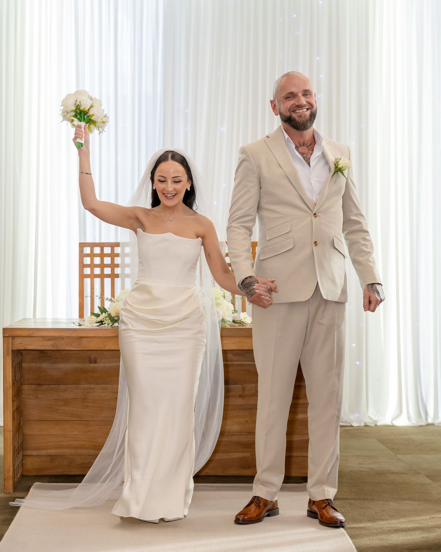 Celebrating newlyweds walk down the aisle, bride raising white bouquet triumphantly after ceremony.