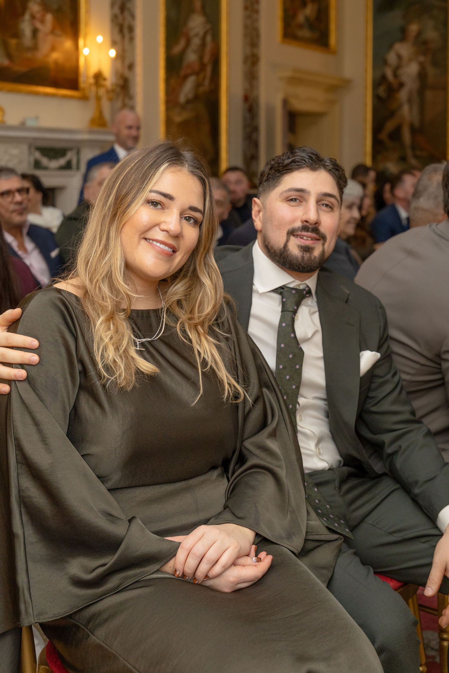 Couple sitting together at formal event, woman in olive dress and man in grey suit with green tie.