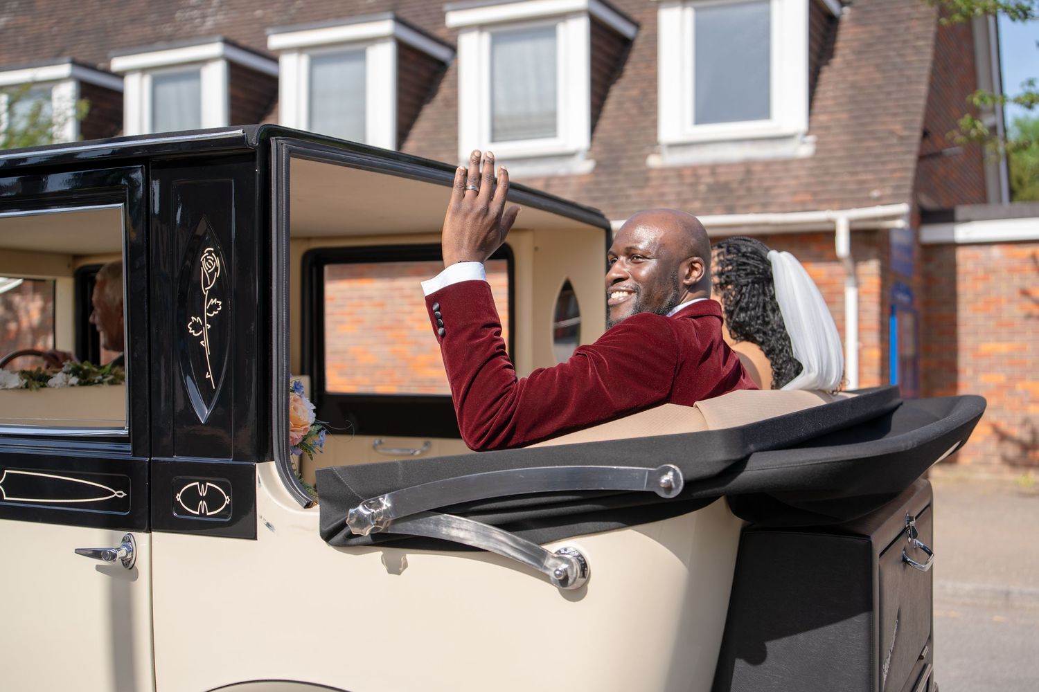 Elegant man in burgundy suit waving from a vintage cream classic car on a sunny residential street.