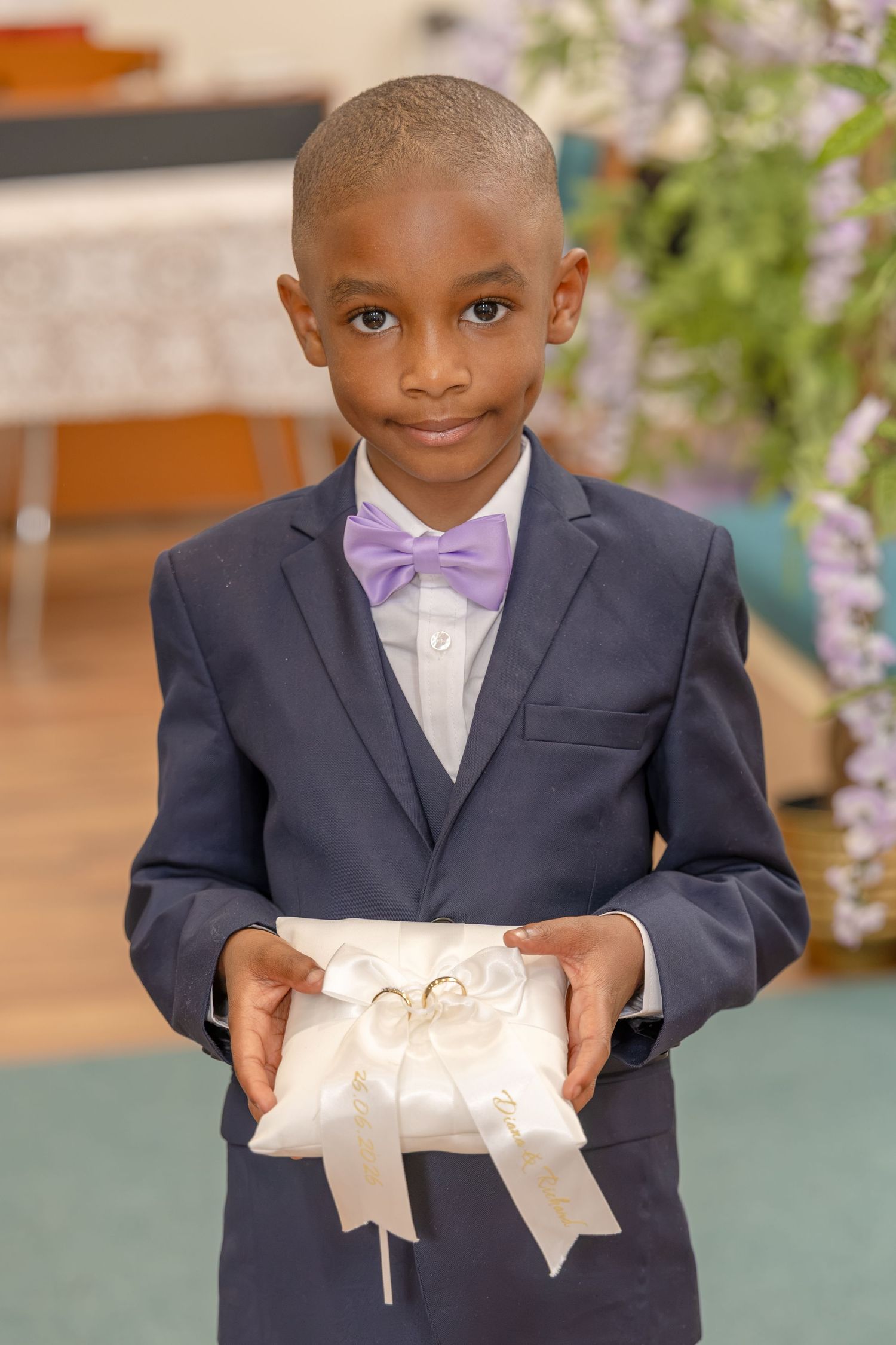 Young ring bearer in navy suit and lavender bow tie holds white pillow with wedding rings at ceremony.