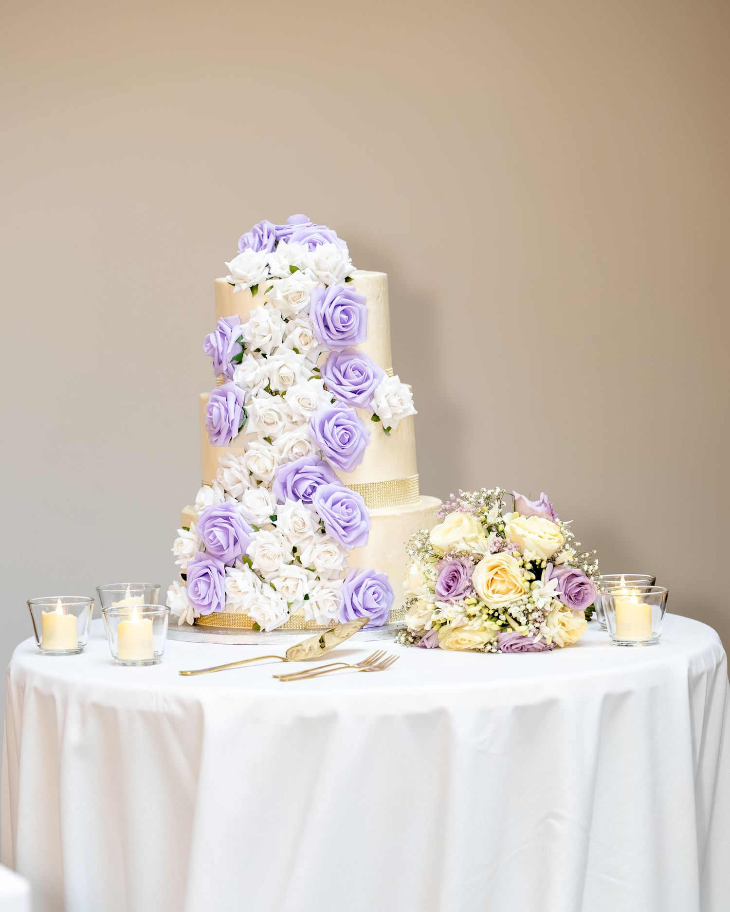 Elegant four-tier wedding cake adorned with cascading purple and white roses on a white draped table with candles.