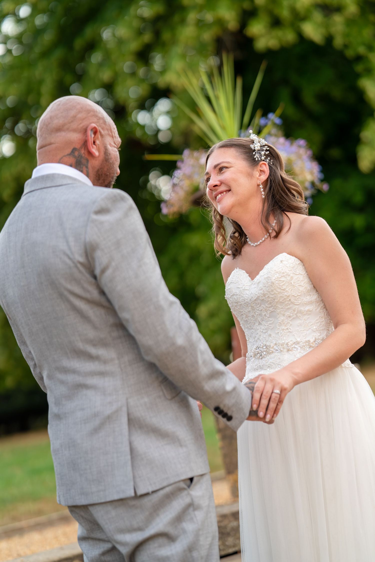 Bride in strapless lace gown smiles at bald groom in gray suit while holding hands outdoors at wedding ceremony.