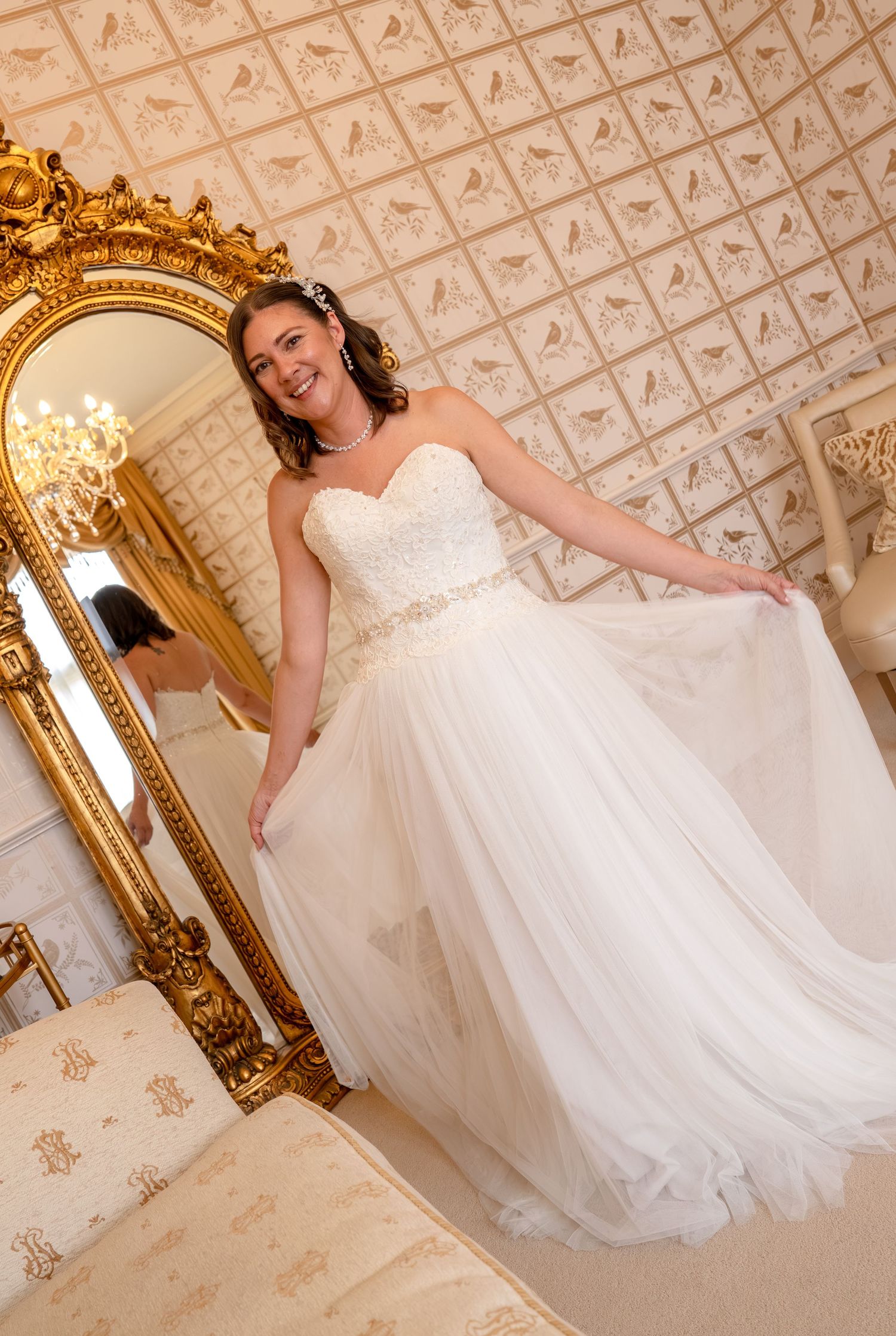 Smiling bride in strapless white tulle wedding gown posing beside ornate gold mirror in elegant room.