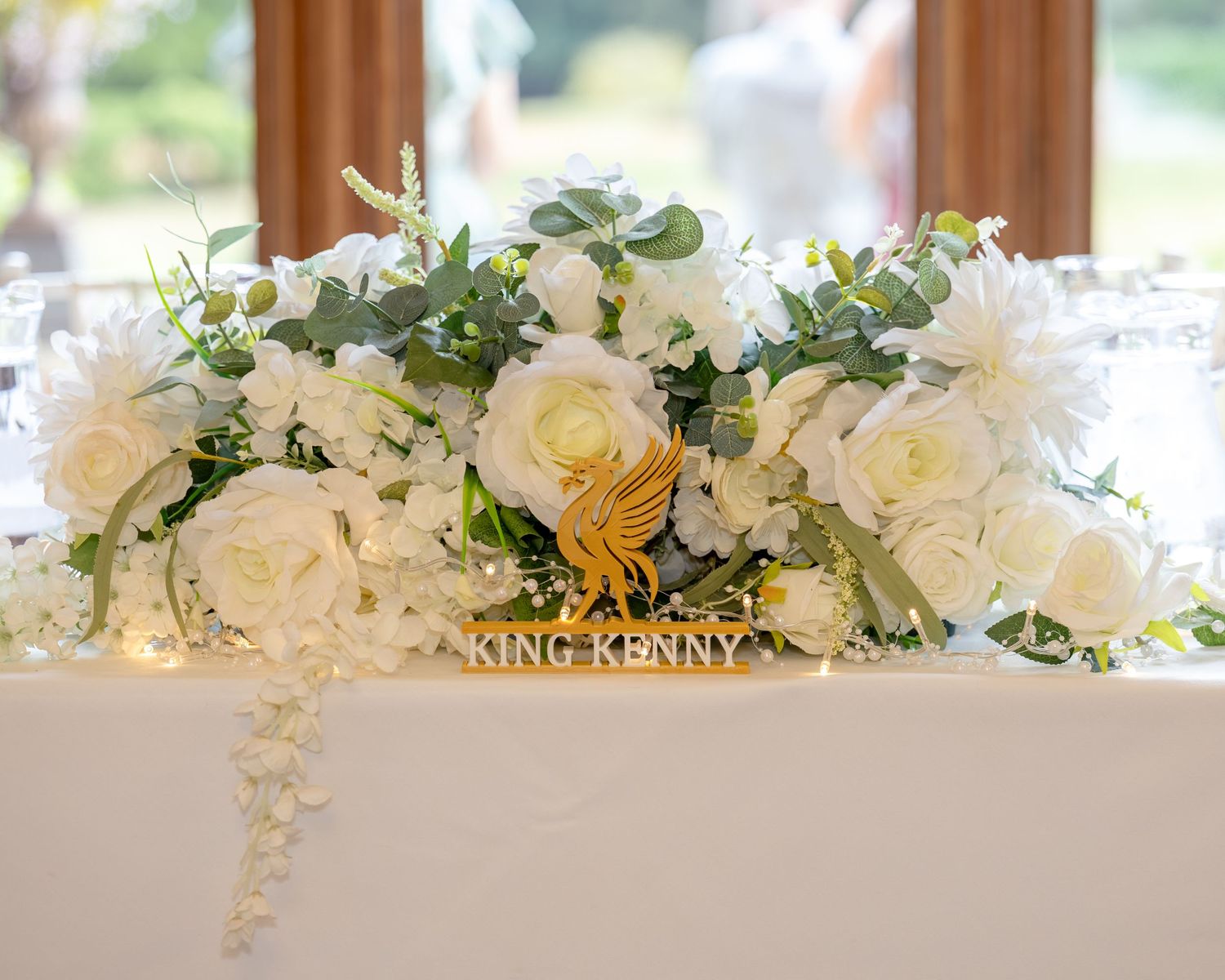 Elegant wedding head table adorned with lush white roses and greenery floral arrangement with a gold King Kenny sign.