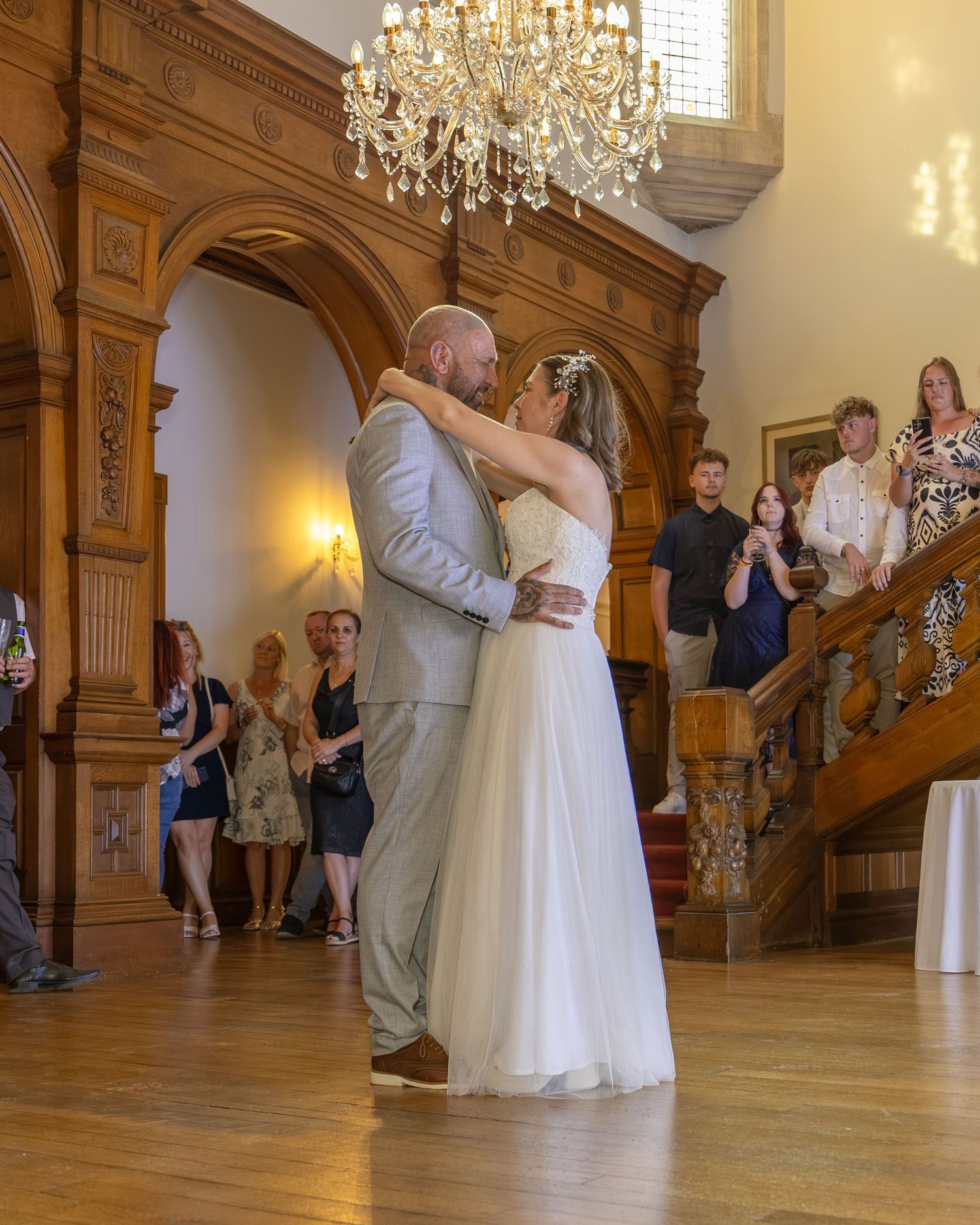 Bride and groom share their first dance in an elegant wood-paneled ballroom with chandelier and guests watching.