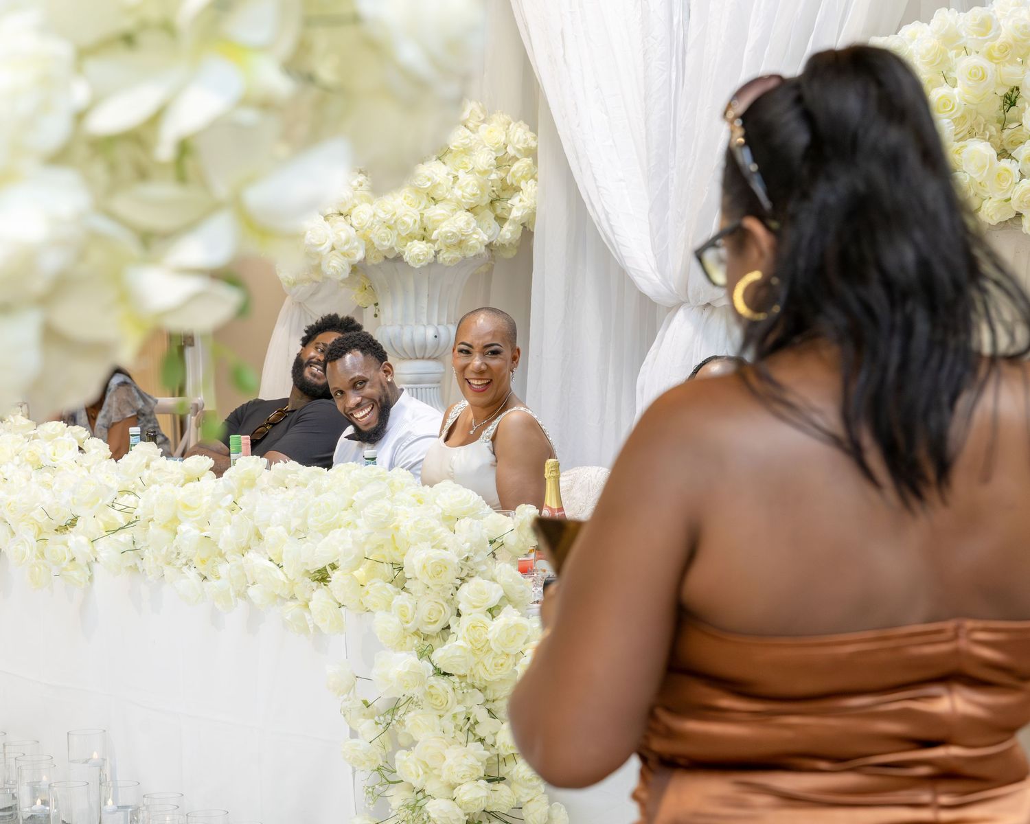 A woman in a bronze dress gives a toast at an elegant wedding reception adorned with white floral arrangements.