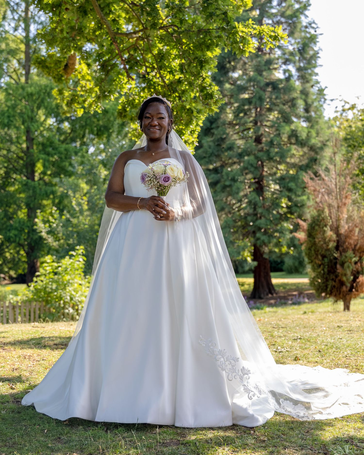 Smiling bride in a white ball gown with veil holds a bouquet outdoors in a lush green garden setting.