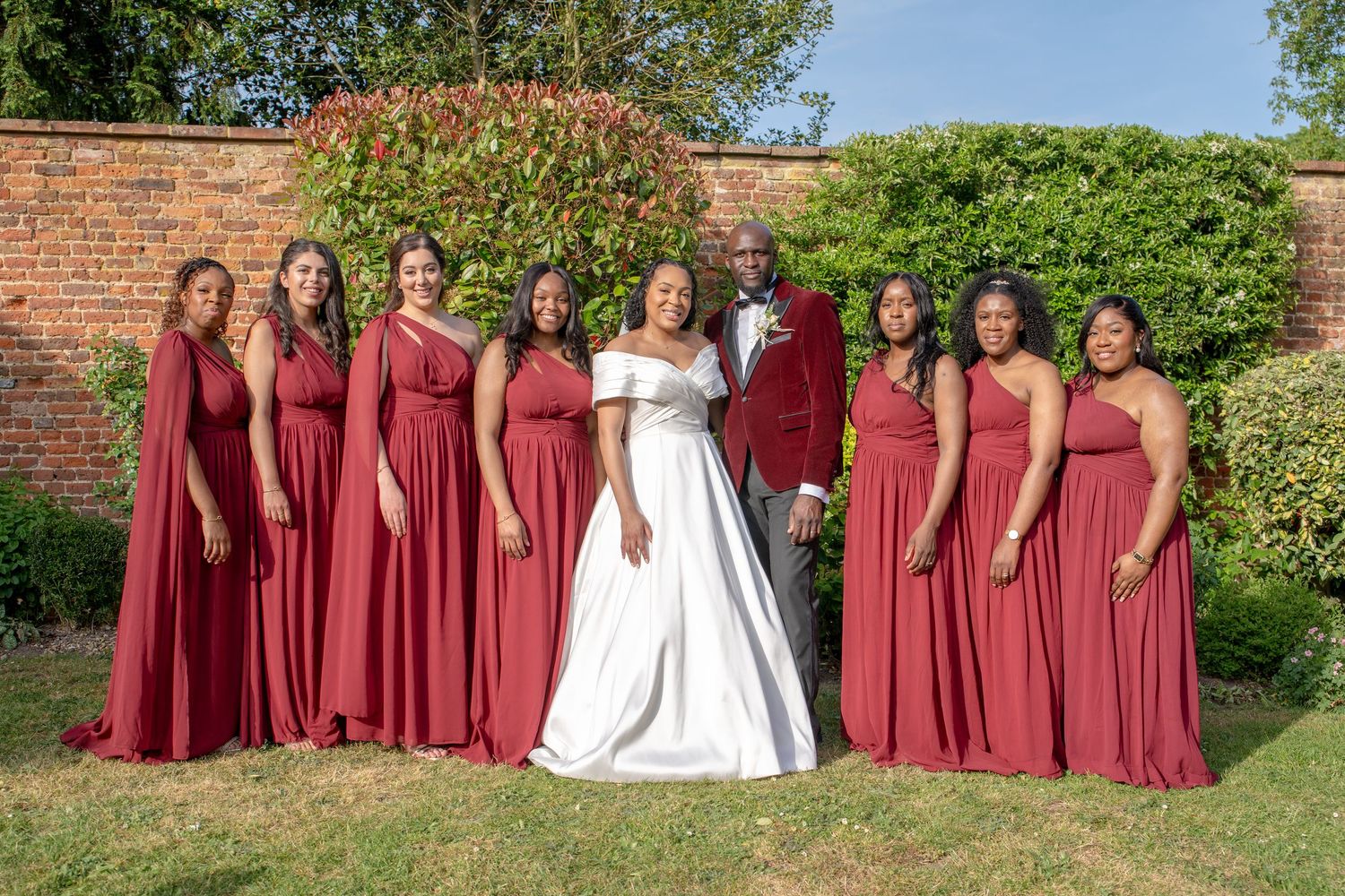 Bride and groom pose with bridesmaids in matching red gowns in a lush garden setting.