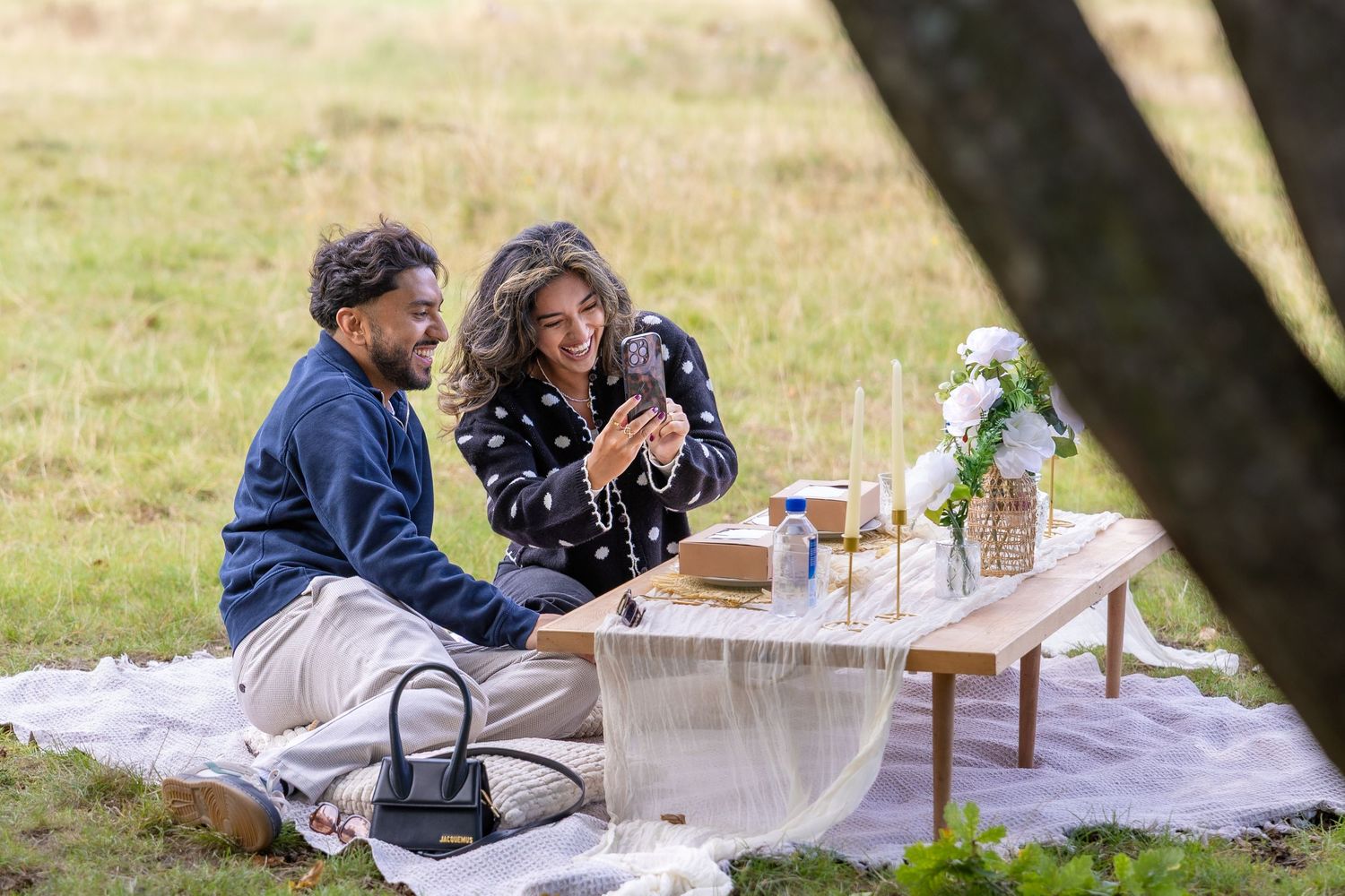 Couple sitting on a picnic blanket looking at engagement ring next to a decorated wooden table outdoors.
