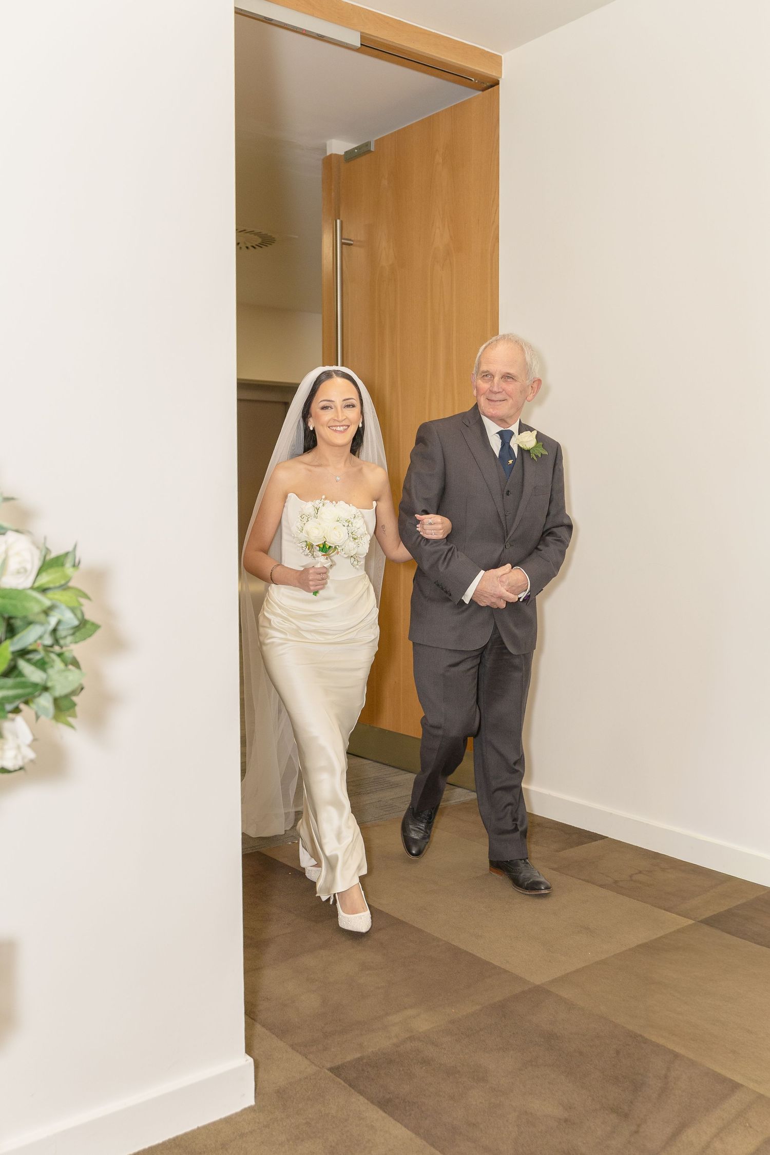 Smiling bride in strapless white gown and veil enters ceremony room escorted by older gentleman in grey suit.