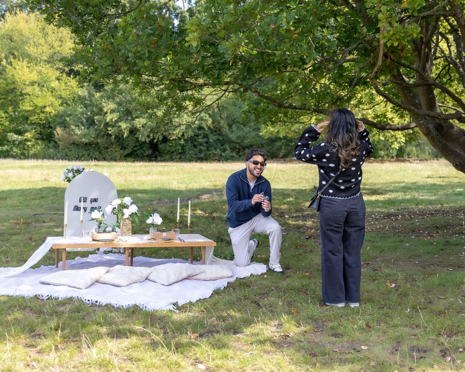 Man on one knee proposing to woman at an elegant outdoor picnic setup in a green park.