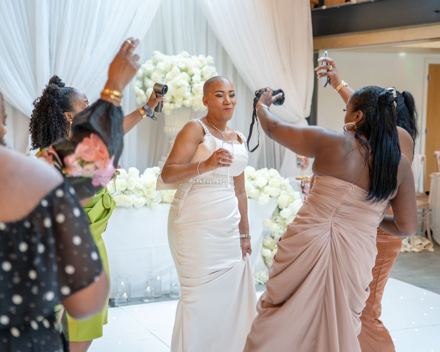 Bride in white gown surrounded by bridesmaids taking photos at elegant wedding reception with floral backdrop.
