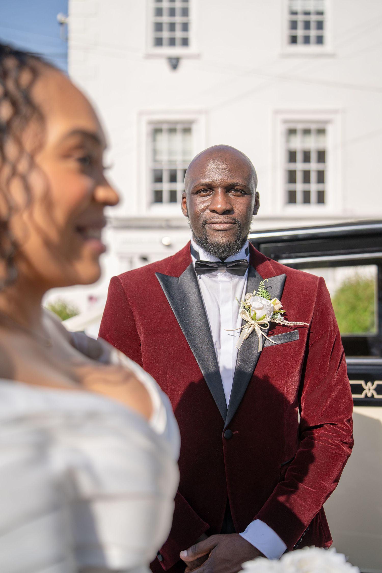Groom in red velvet tuxedo with boutonniere smiles at bride in white dress outside white building.