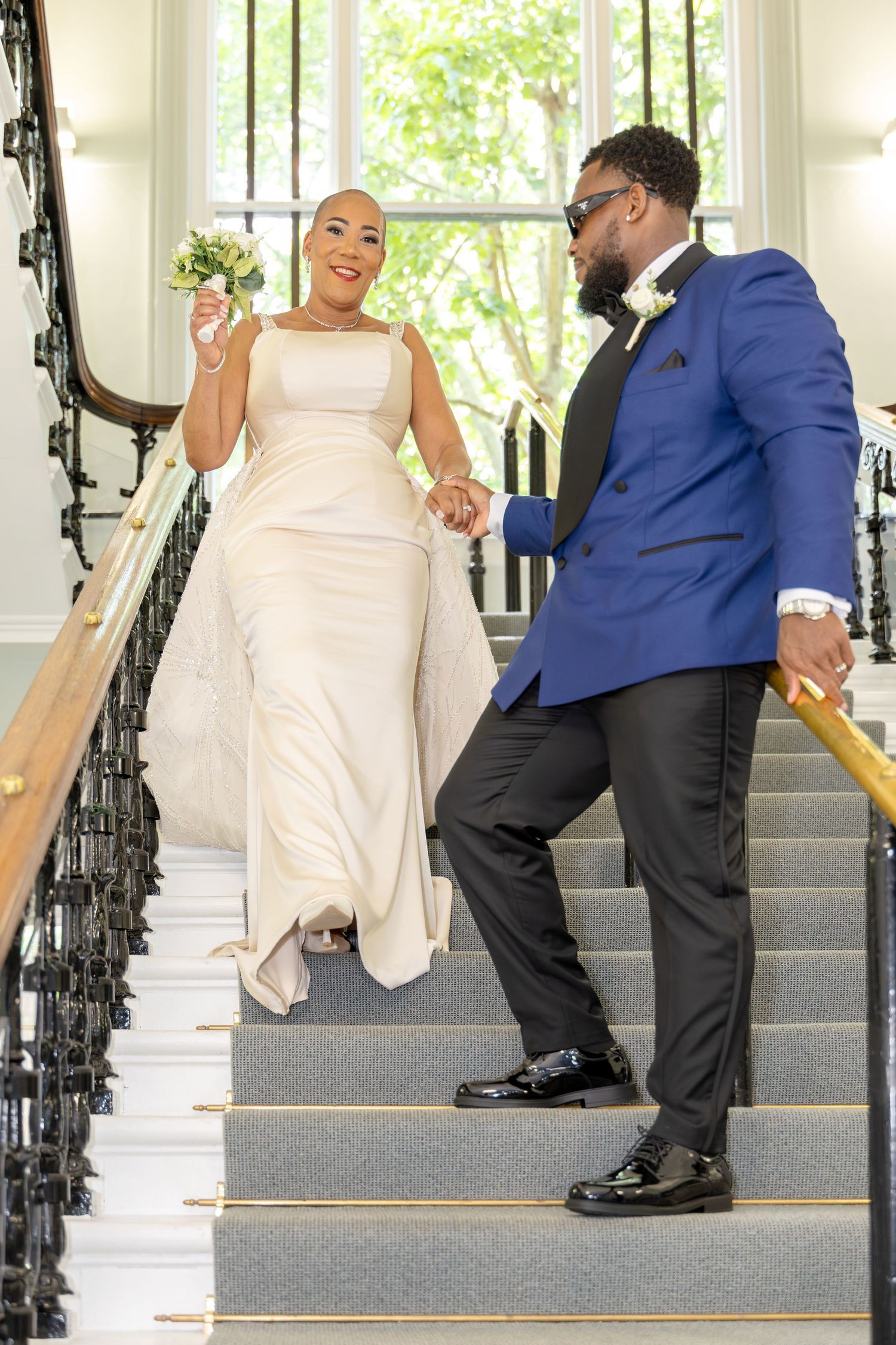 A bride in white gown and groom in blue tuxedo hold hands walking down an elegant staircase on their wedding day.
