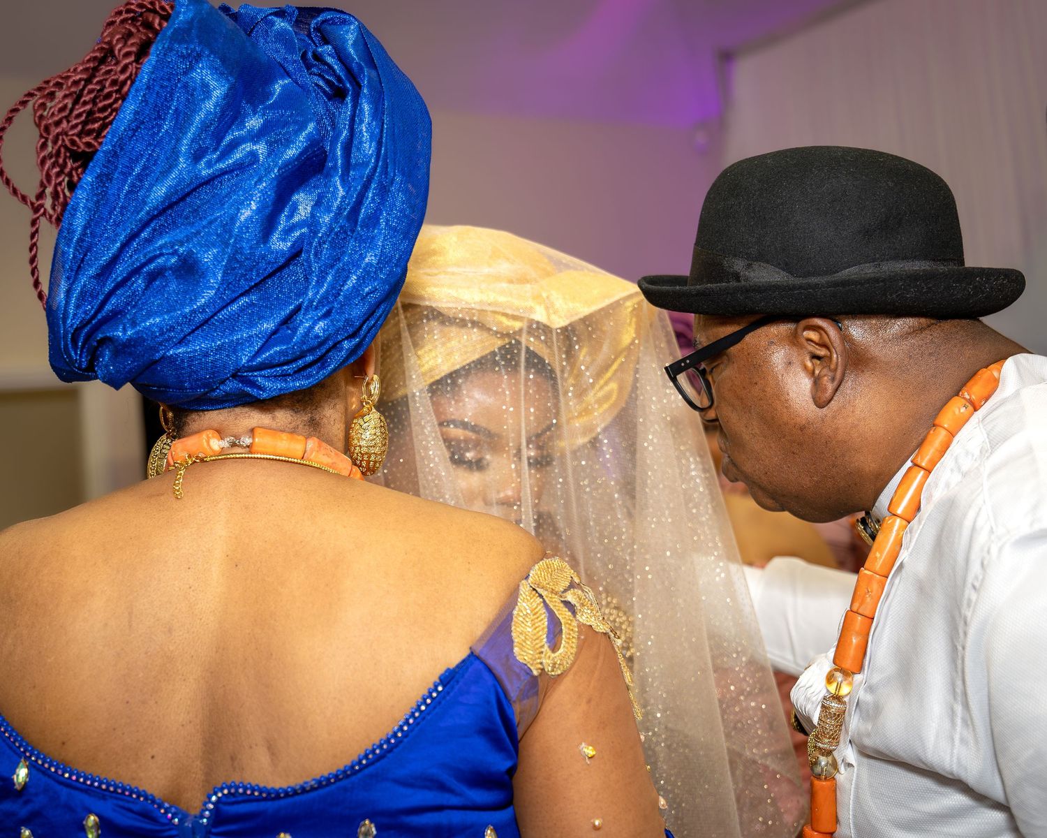 Nigerian bride in gold gele receives blessing from elders in blue aso-oke at traditional wedding ceremony.