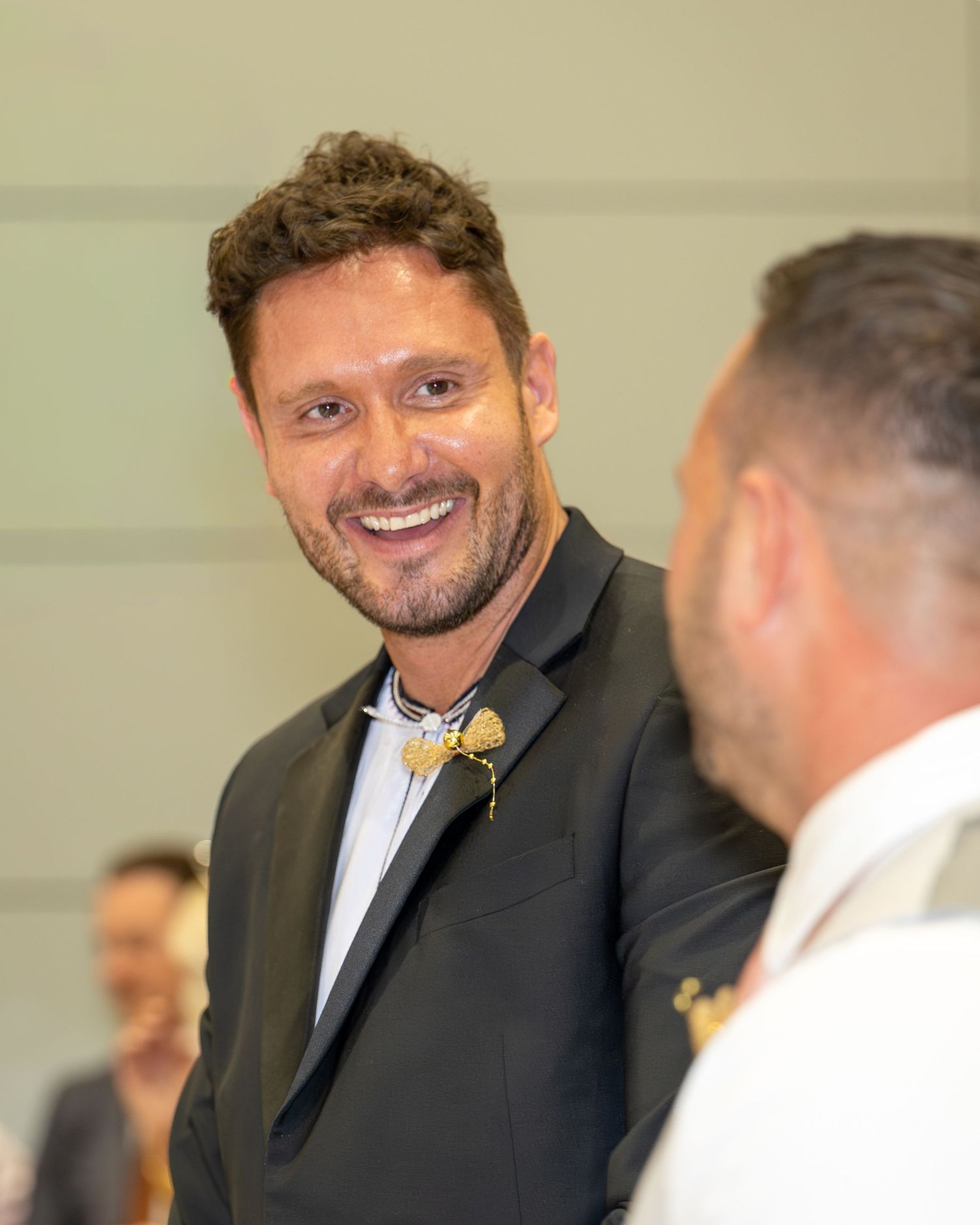 Smiling groom in black suit with gold boutonniere at a wedding reception.