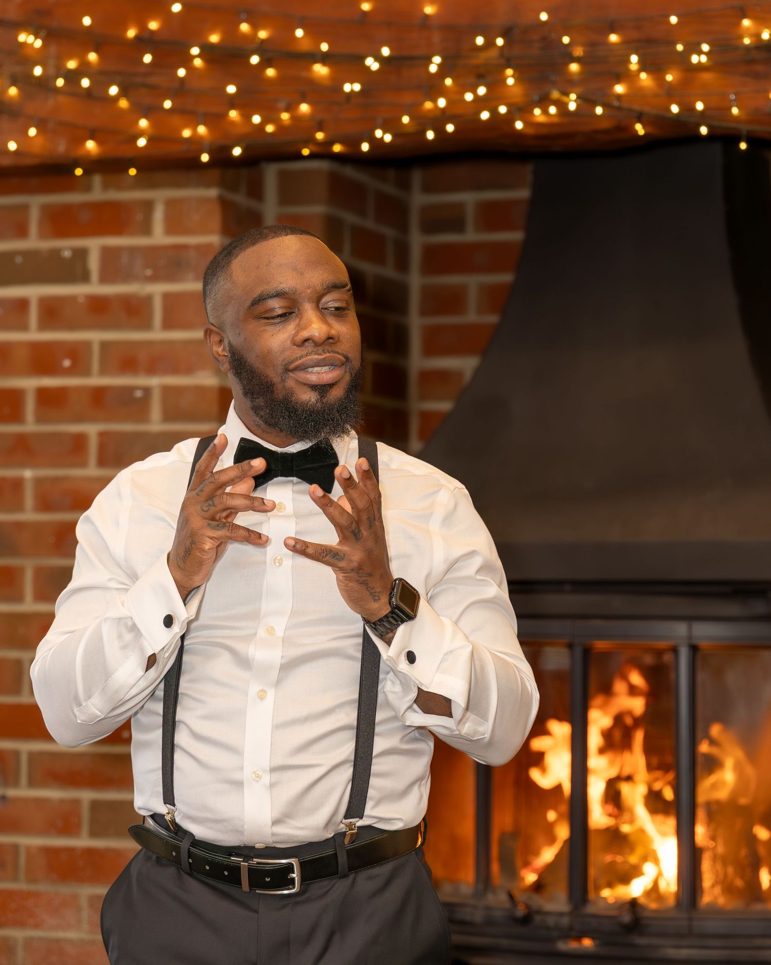 Groom in white shirt, suspenders, and bow tie poses by a fireplace with fairy lights at a wedding reception.