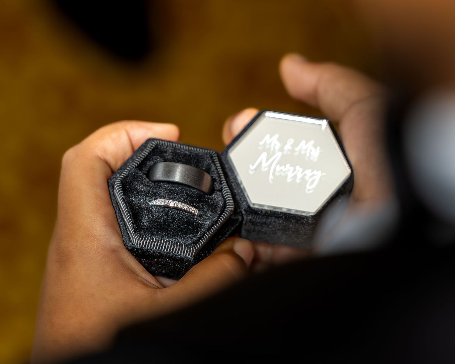 Hands holding open a black hexagonal ring box with white interior revealing an engagement ring.