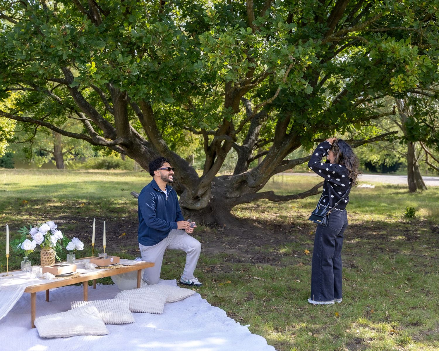 Man kneeling on a white blanket proposing to woman at a romantic outdoor picnic in a park.