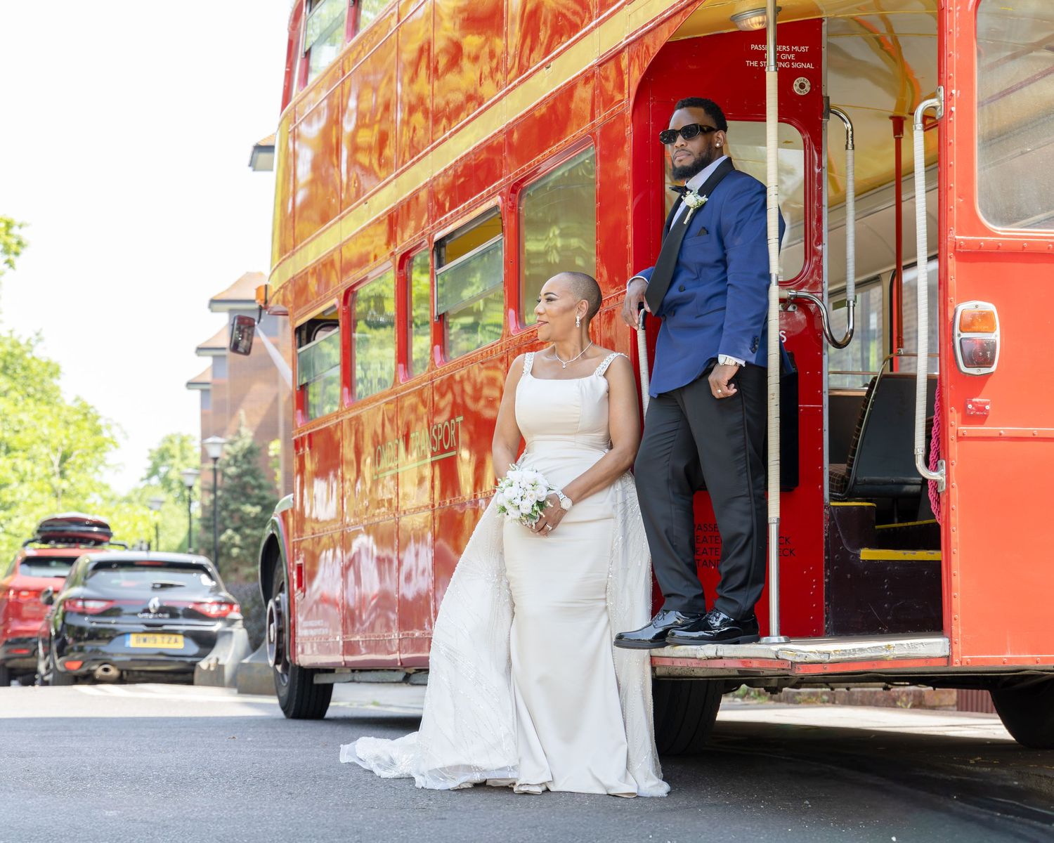 Bride in white gown and groom in blue suit pose by a red double-decker bus on their wedding day.