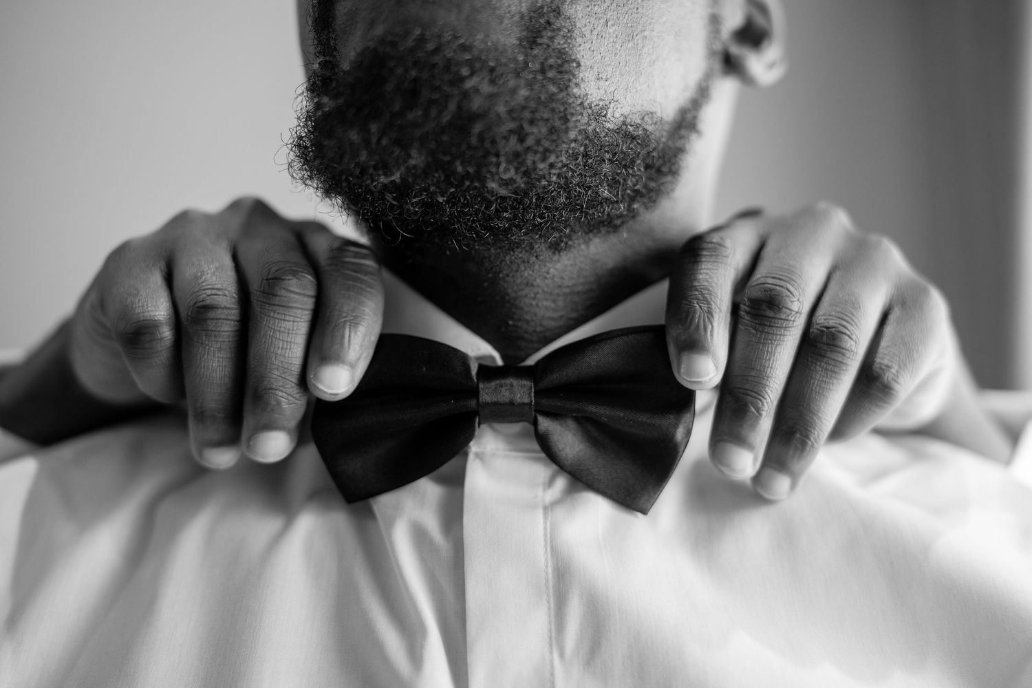 Black and white photo of a man adjusting his bow tie, wearing a white dress shirt, with a neatly groomed beard.