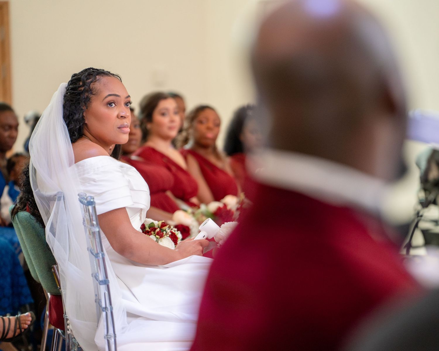 Bride in white veil seated at wedding ceremony with bridesmaids in red dresses behind her.