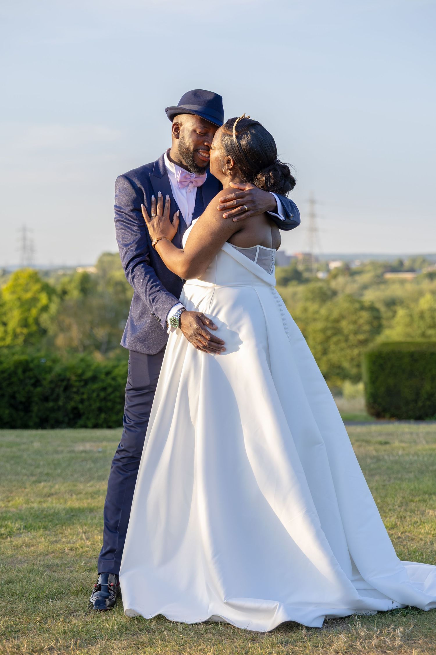 A groom in a navy suit and hat kisses his bride in a white ballgown outdoors on their wedding day.