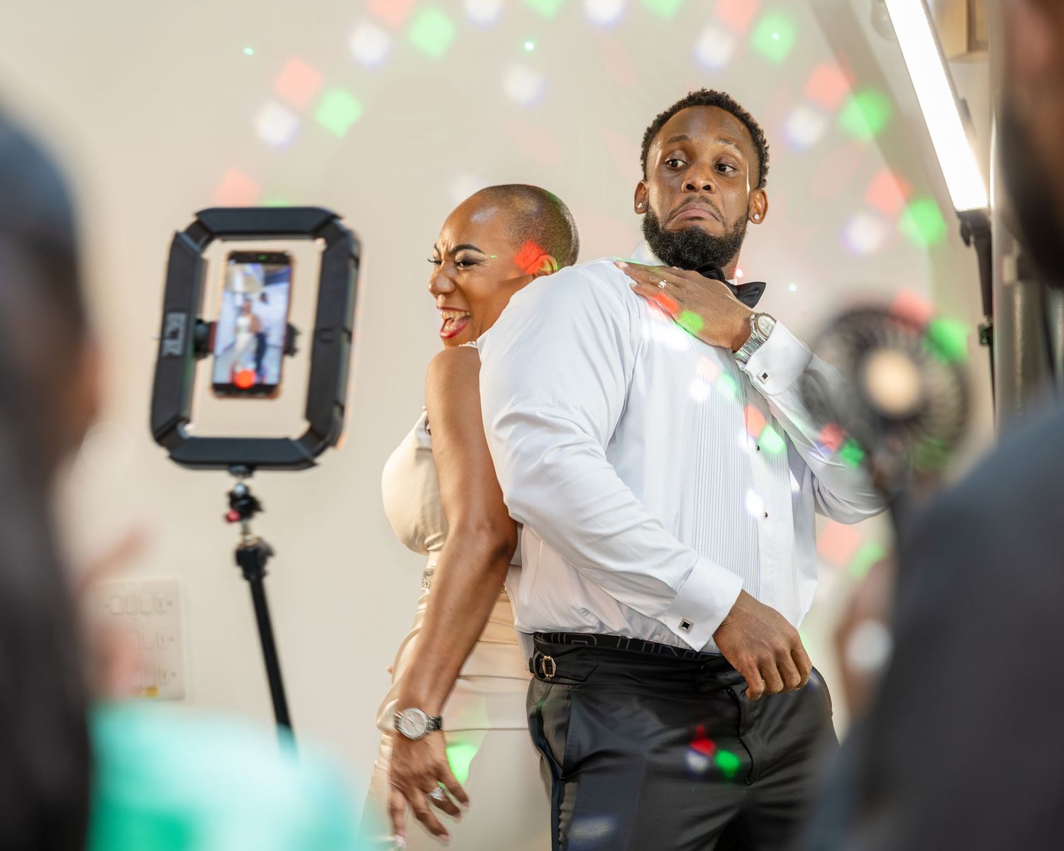 Couple posing back-to-back at a party with colorful disco lights and a phone on a ring light stand.
