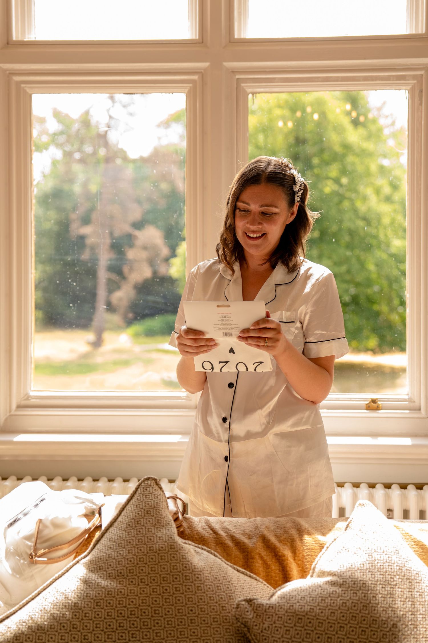 A smiling woman in white pajamas reads a card by a sunlit window with a lush garden view.
