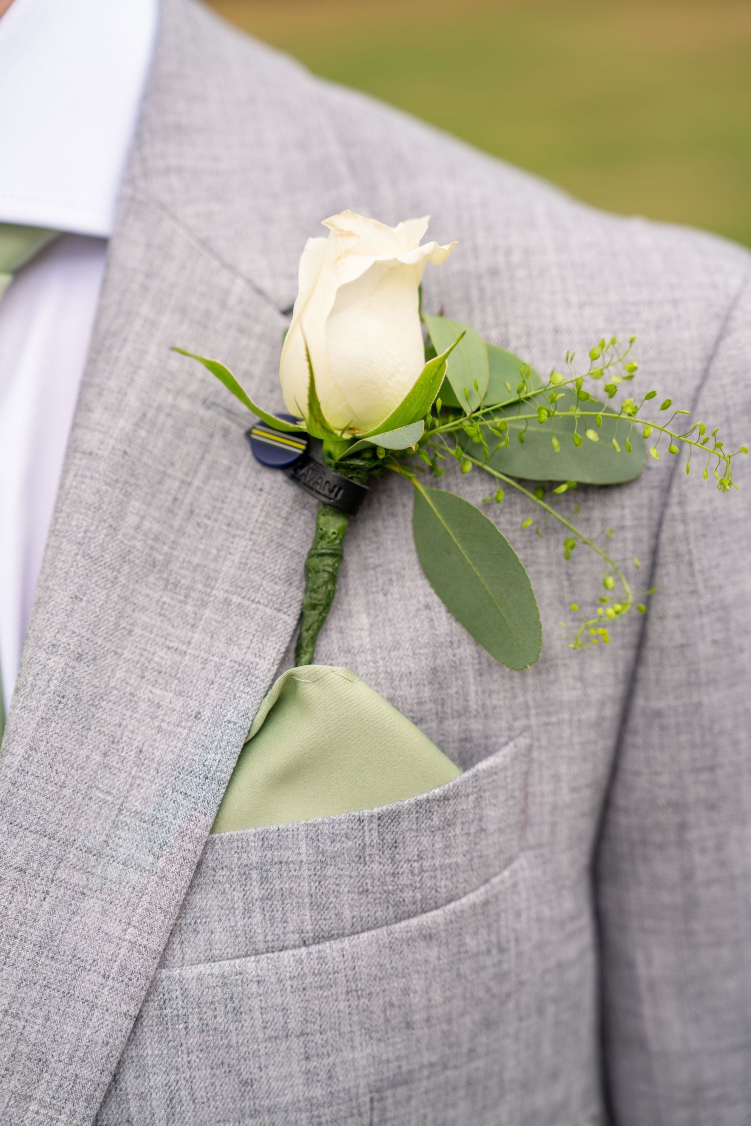 Gray suit with white rose boutonniere and sage green pocket square, perfect for a wedding or formal event.