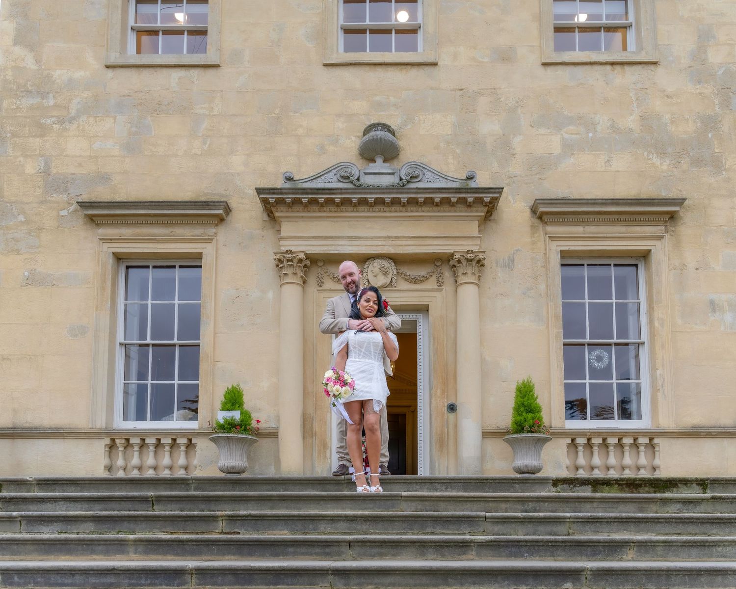 Groom lifts bride on grand stone staircase outside elegant manor house wedding venue.