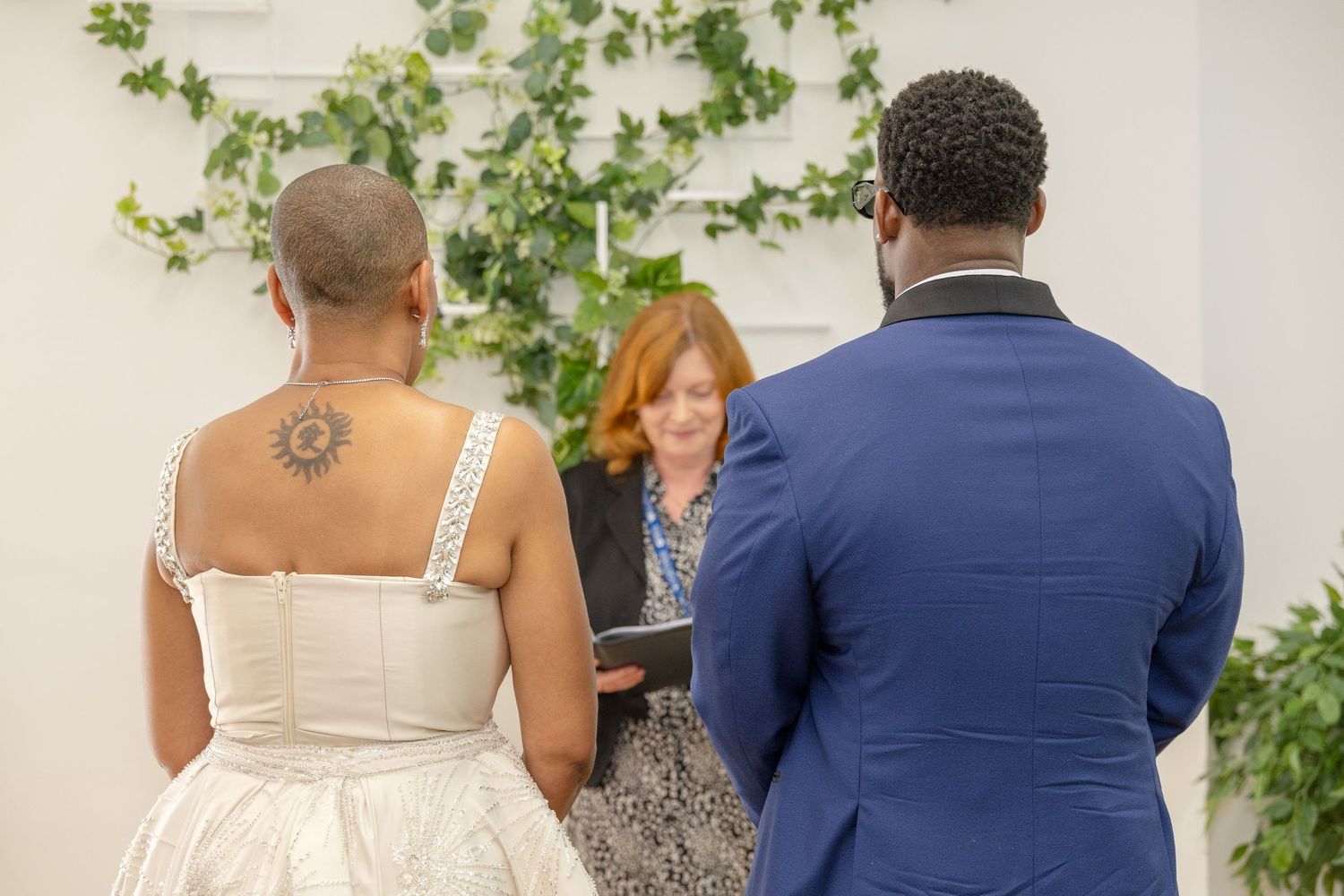 Couple exchanging vows at wedding ceremony with officiant holding book, bride in white dress with tattoo, groom in blue suit.